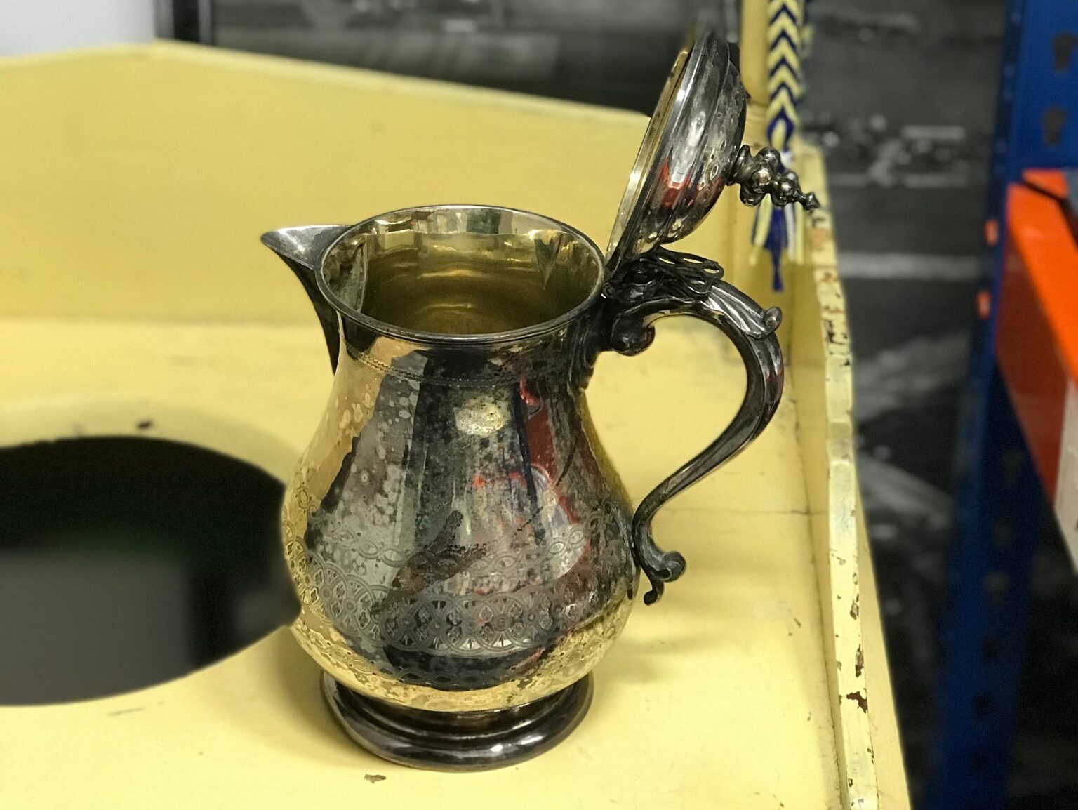 Silver plate jug with an ornate handle and hinged lid, displayed on a yellow wooden stand.