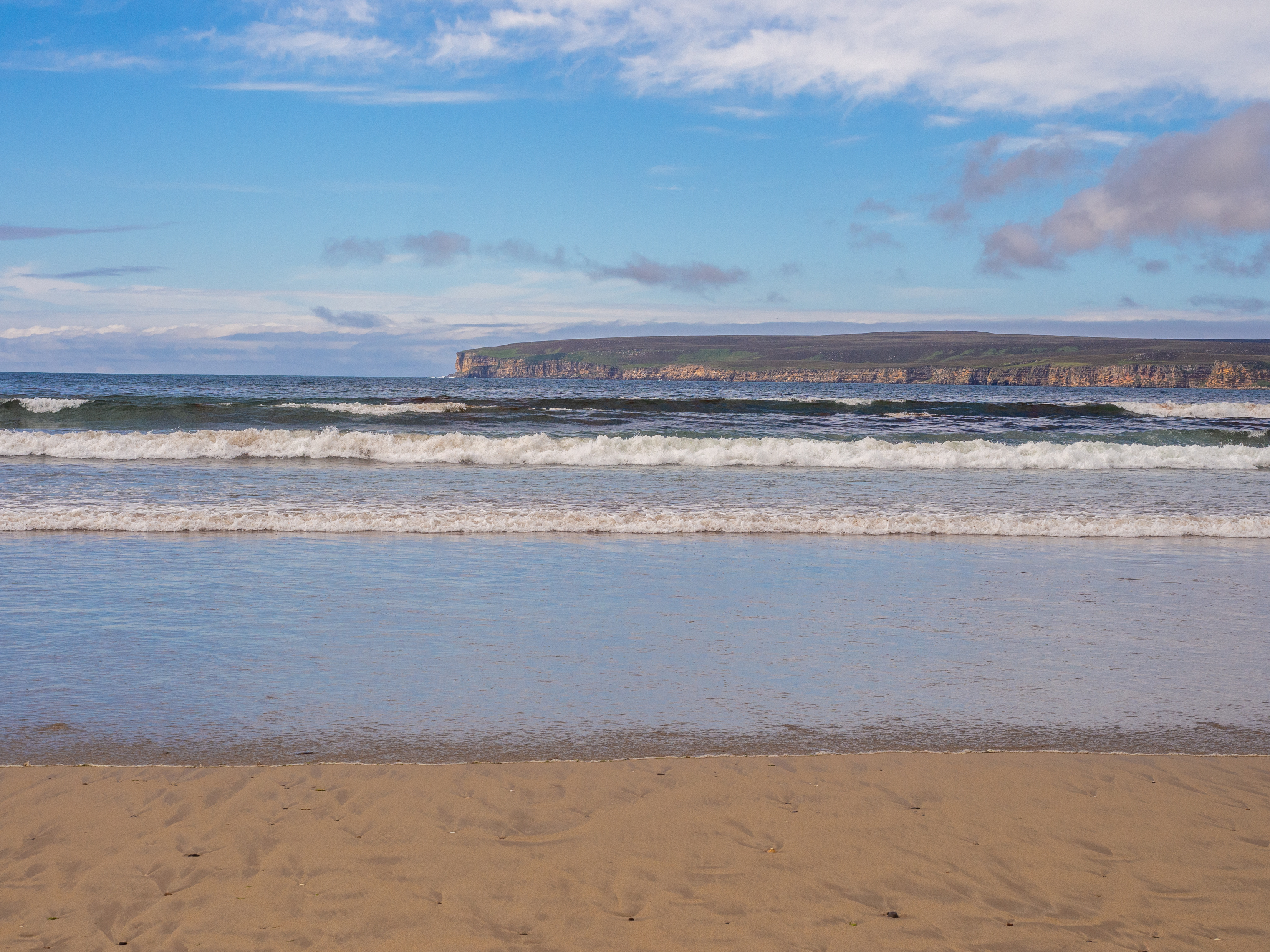 Gentle waves rolling onto a sandy beach with cliffs in the distance.