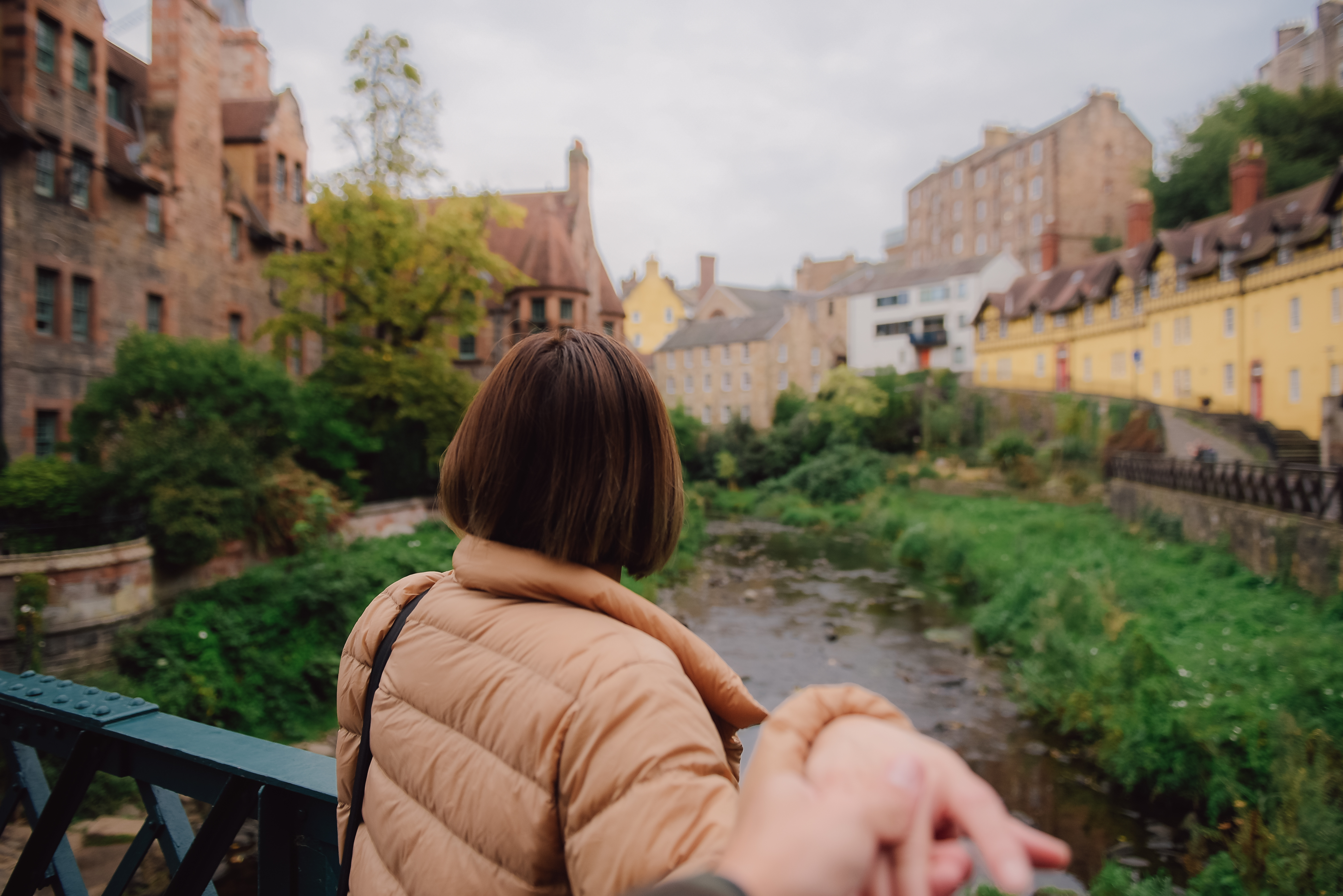 A person in a tan jacket holding someone’s hand while looking over a leafy riverside area.