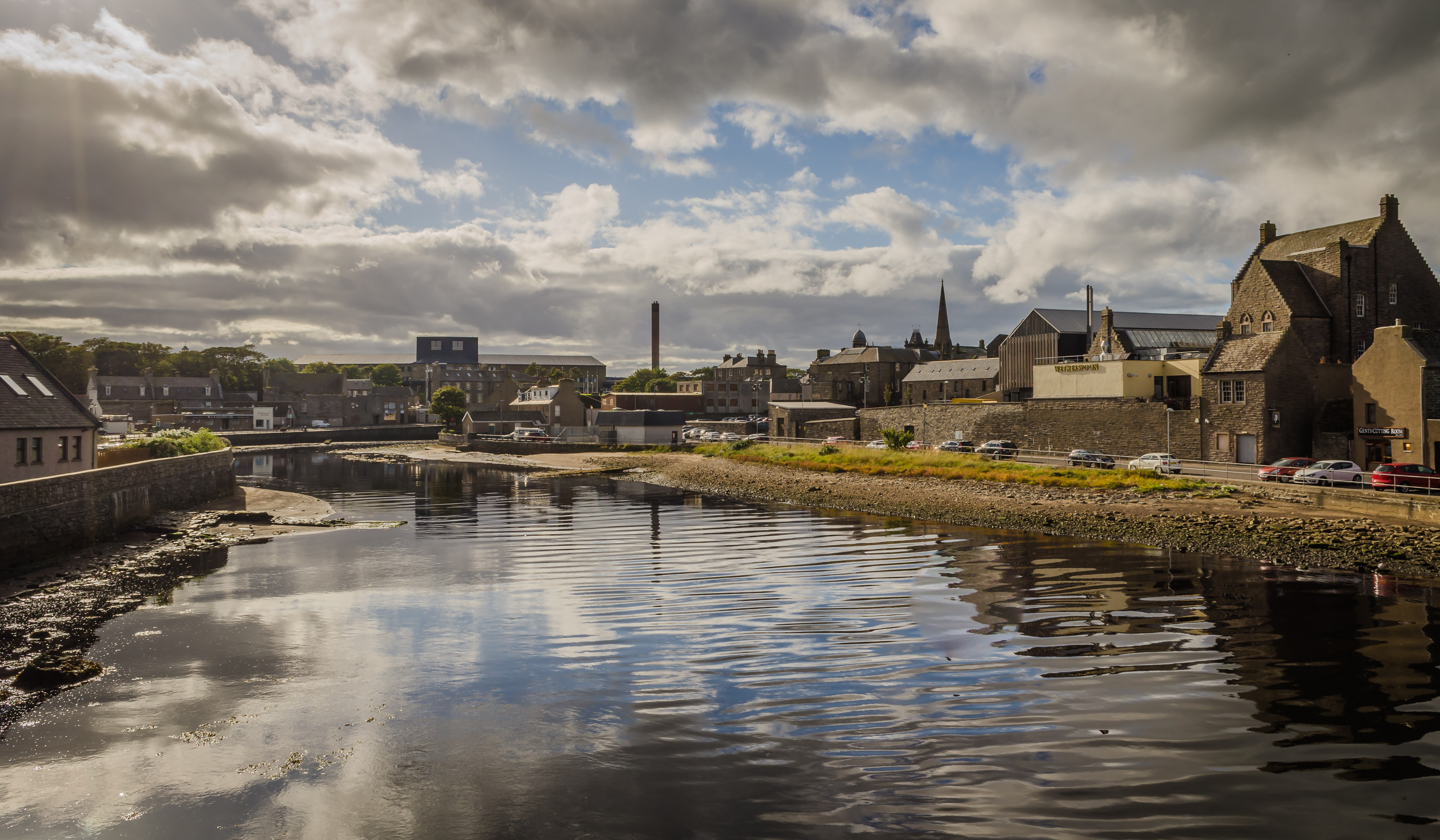 A riverside town with reflections of buildings and clouds on the water.