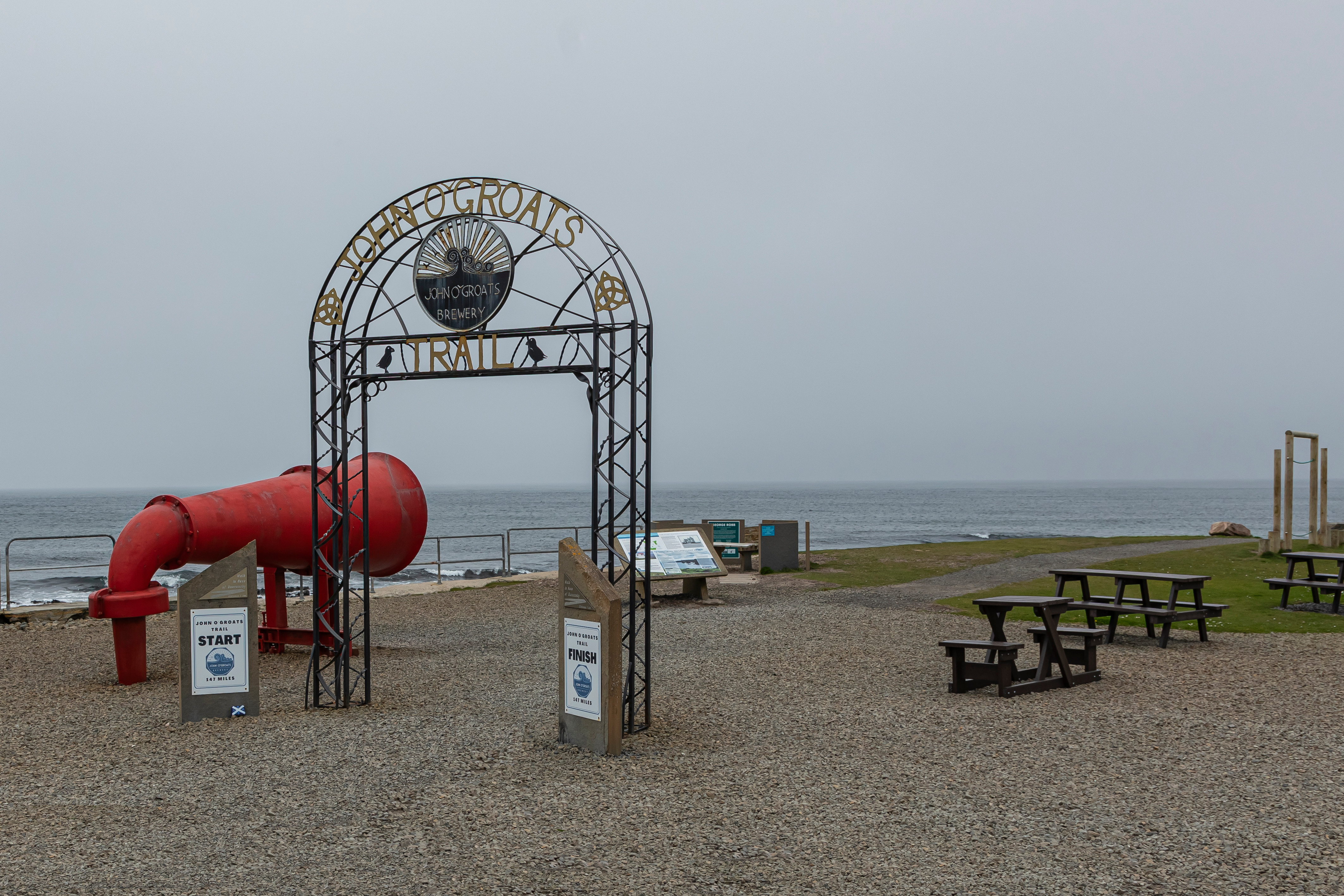 An archway sign marking the start of a coastal trail near picnic benches and the sea.