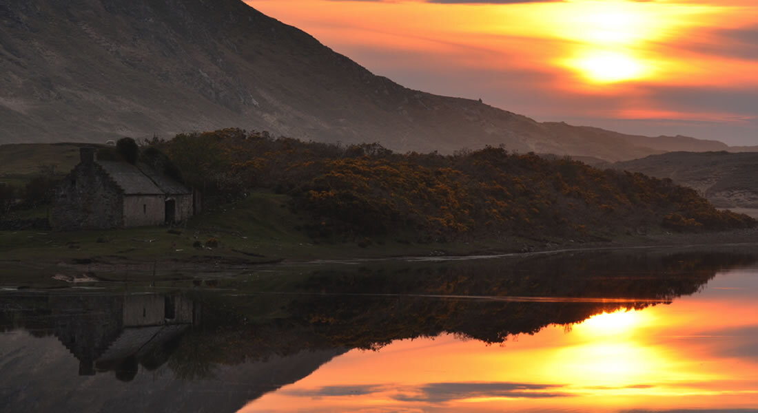 A sunset over hills and water with an old stone cottage silhouetted in the foreground.