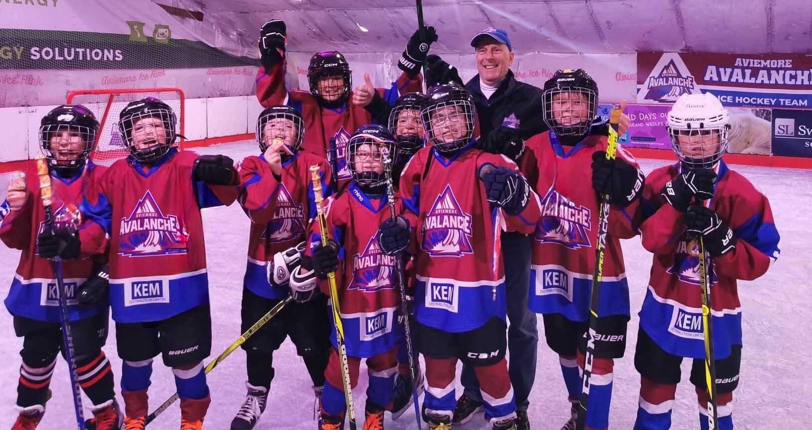 A group of nine young ice hockey players in red and blue hockey tops. They are wearing helmets and holding up ice hockey sticks. They are in an ice rink.