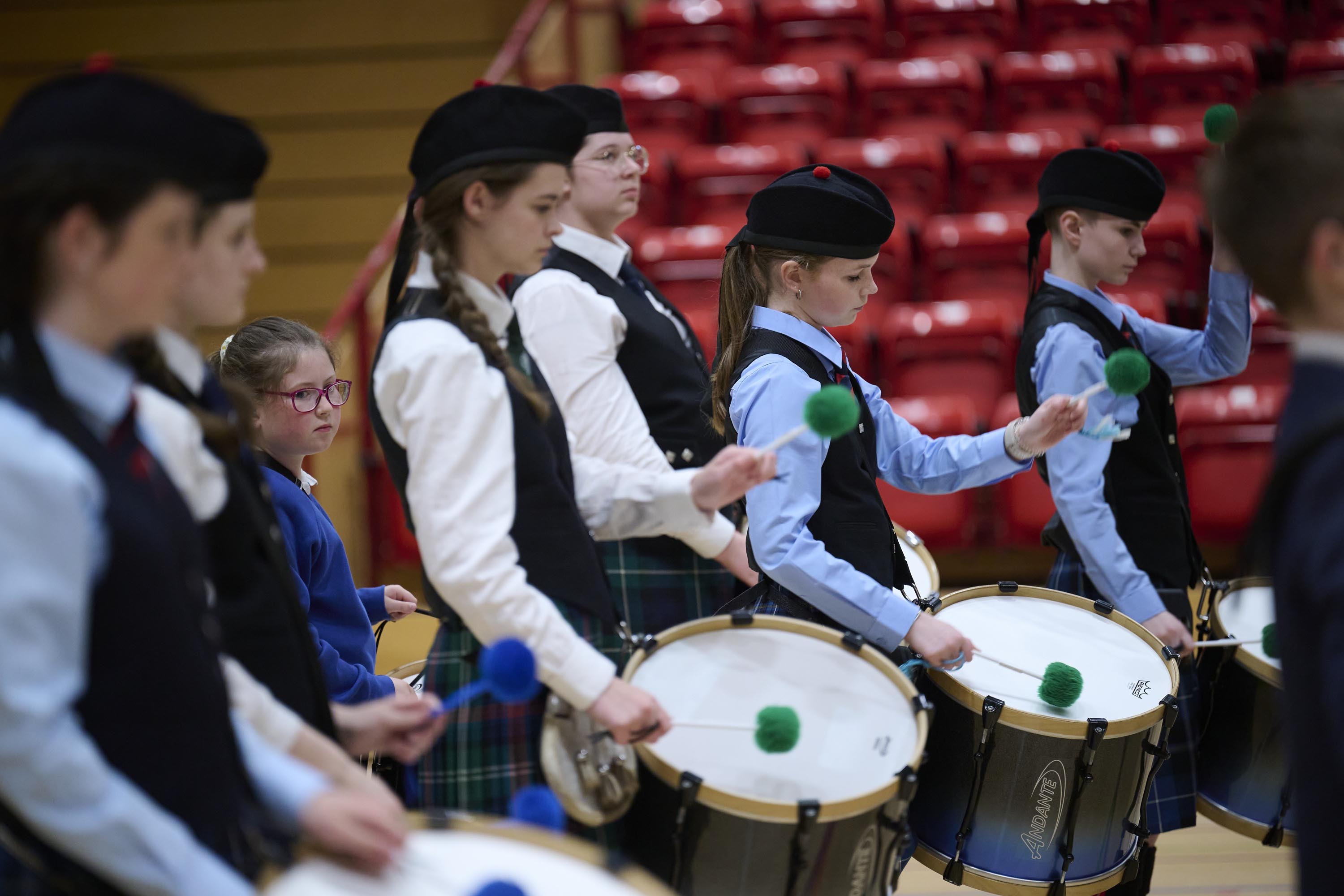 A group of young people playing the drums and marching in a gym hall setting. They are wearing dark blue kilts, pale shirts and black waistcoats.