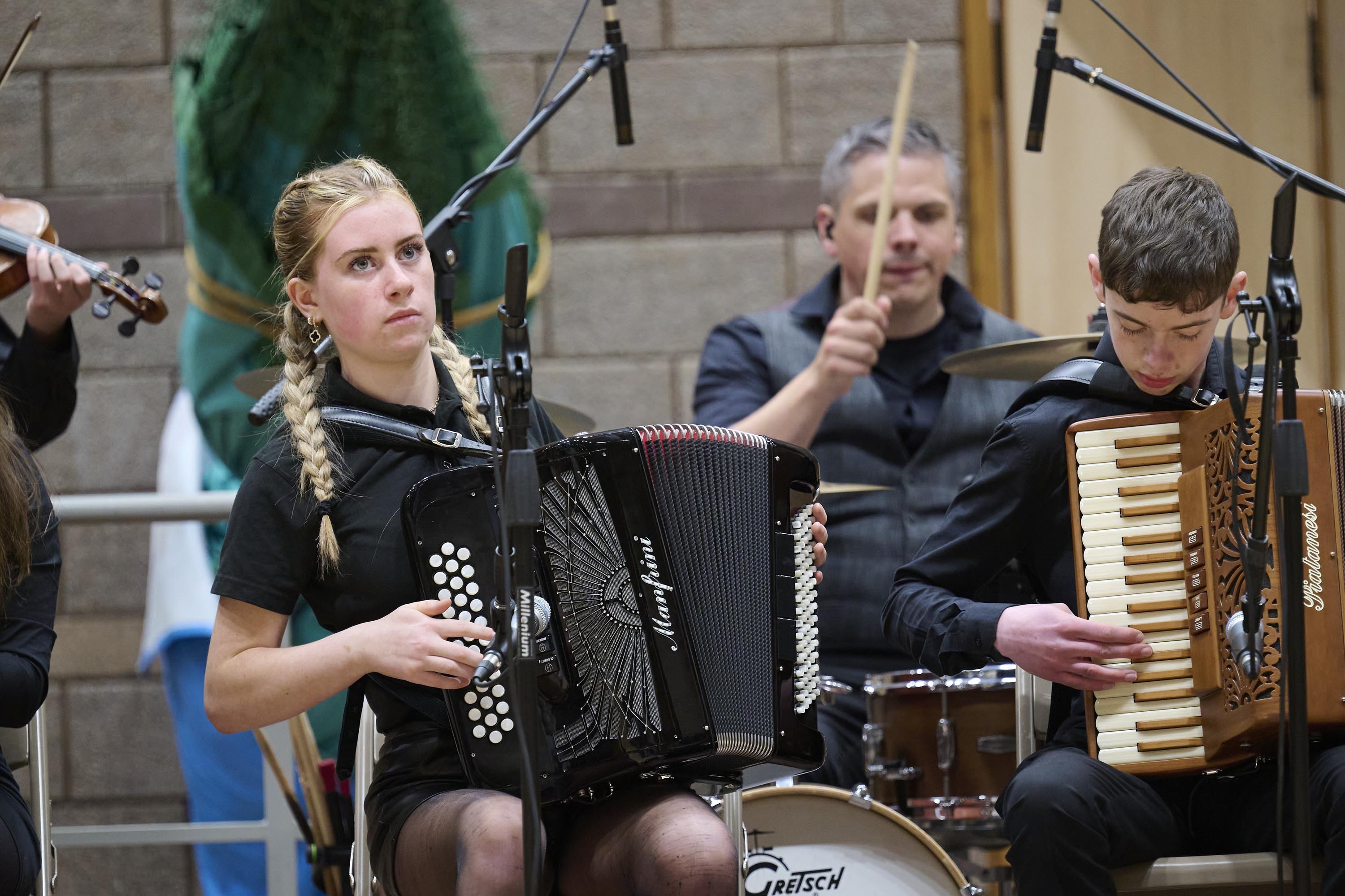 Two young people - a female on the left and a boy on the right - are seated and playing the accordion. Behind them is a man playing a drum kit.