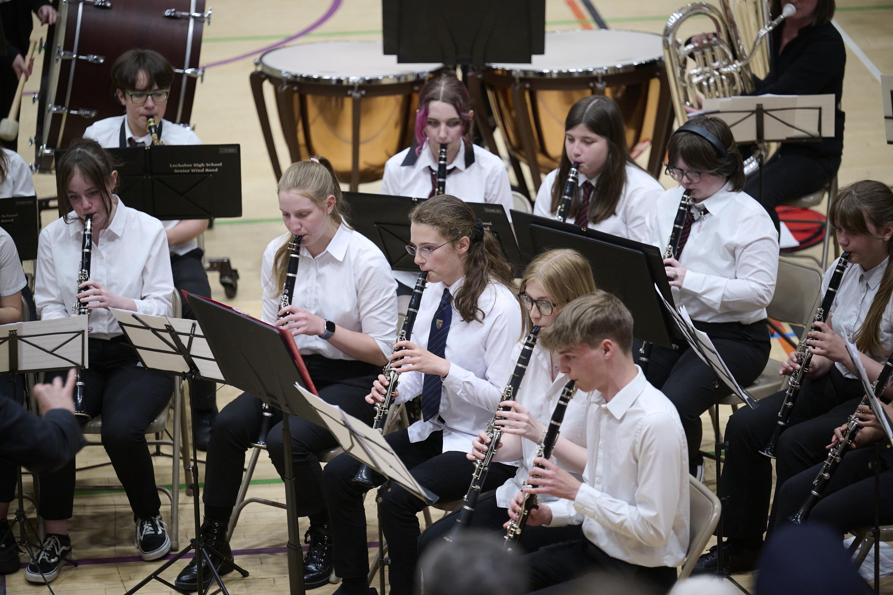 A group of young people are seated and playing the clarinet. They all have music stands in front of them. They are wearing white shirts.