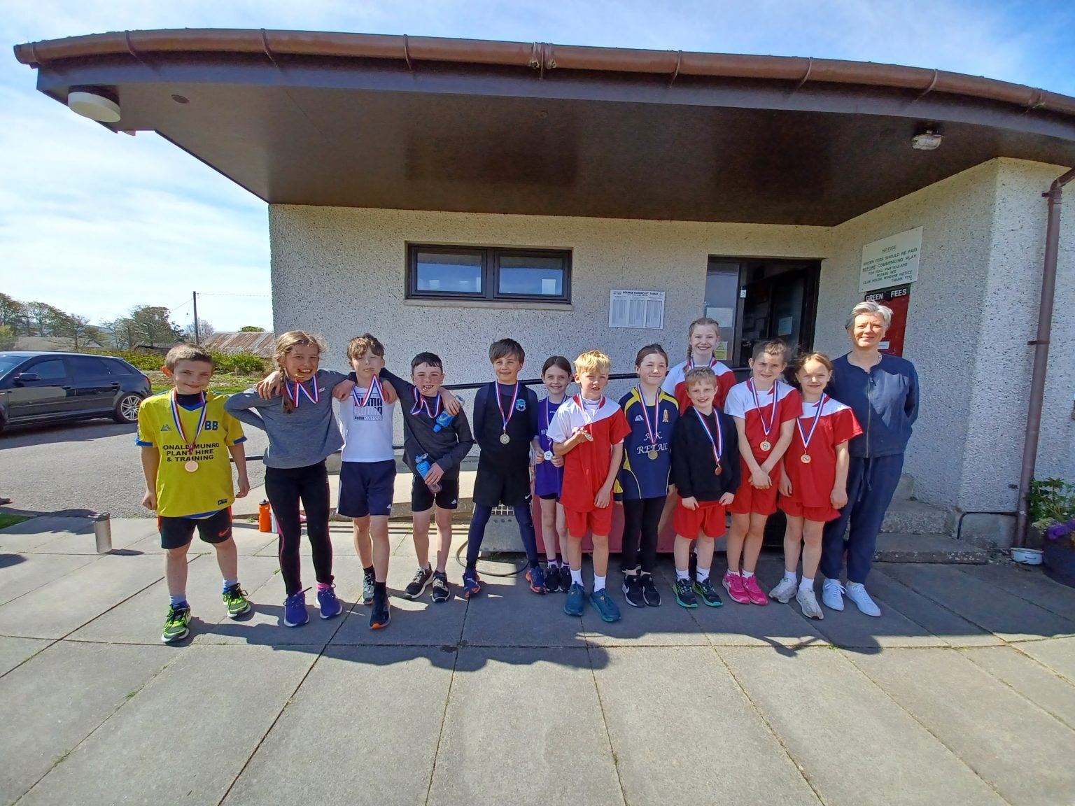 A group of children standing in a row outside a light-colored building with a dark roof. They are wearing different sports outfits and each child has a medal around their neck