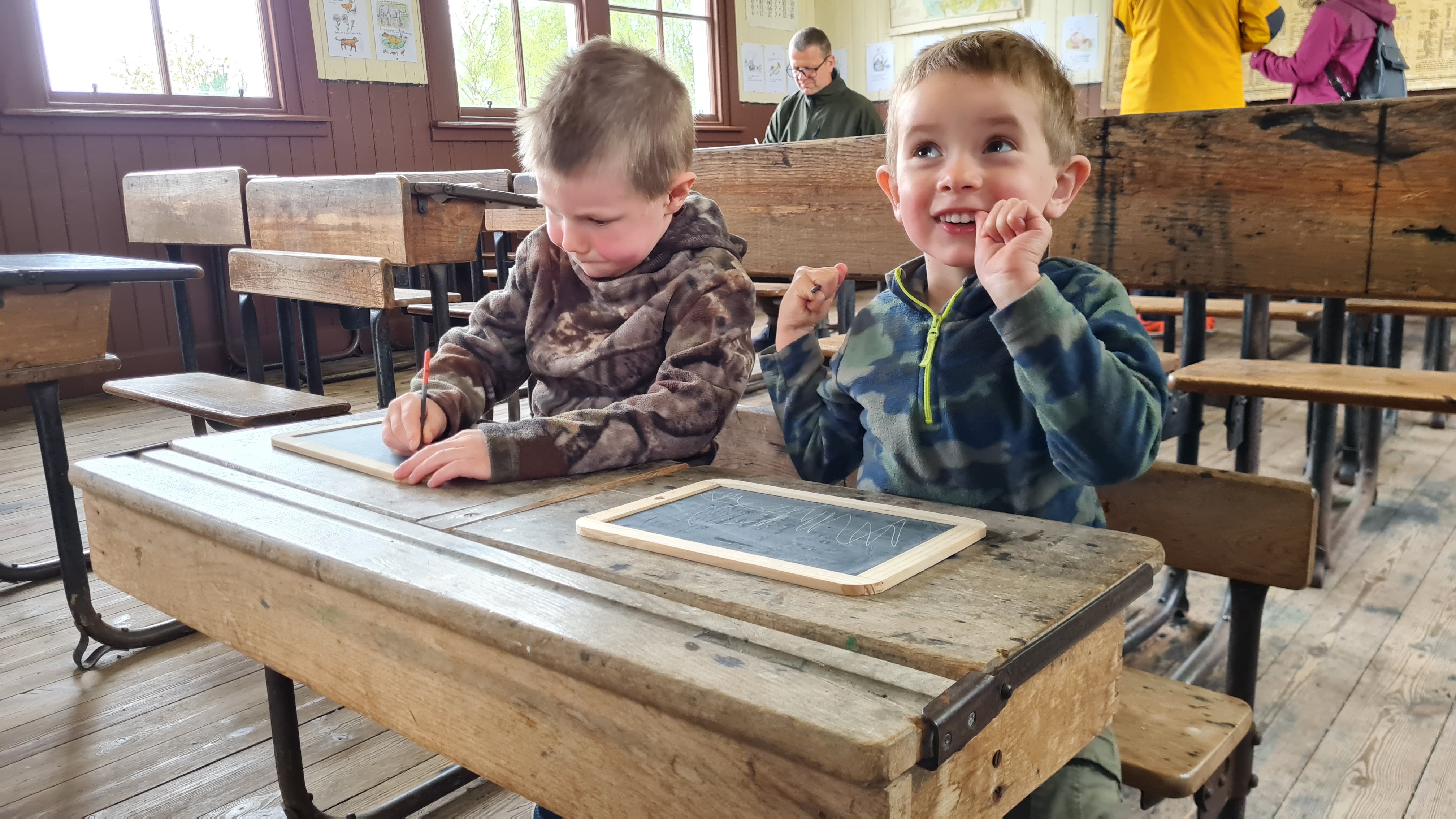 Two young children sit together at an antique wooden double school desk in a historic one-room schoolhouse. One child writes with a pencil while the other smiles cheerfully at the camera, resting their chin on their hand. A small slate writing board sits on the desk. Rows of period school desks and wooden plank floors are visible in the background.