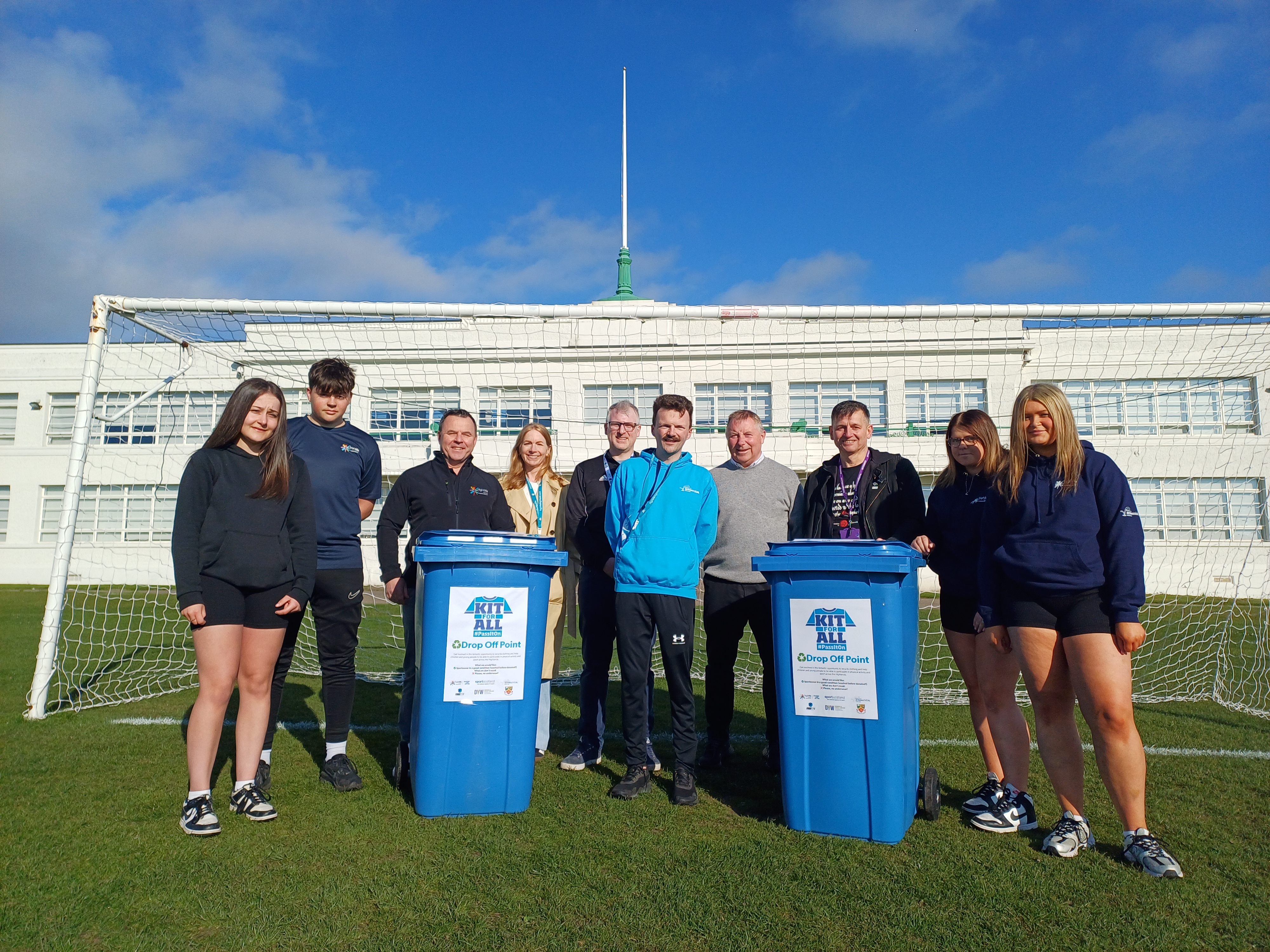Ten people, including four senior school pupils are stood in front of a white building on a sports field. In front of them are two blue wheelie bins with Kit For All branding on them.