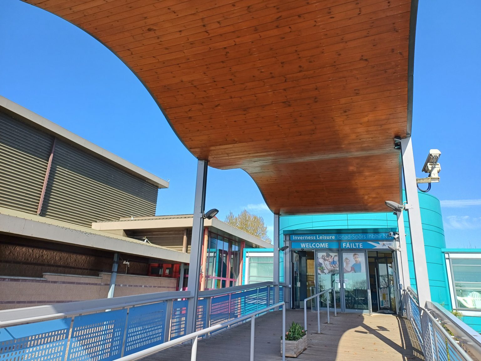 Entrance to Inverness Leisure featuring a modern design with a large curved wooden canopy overhead. A ramp with metal railings leads up to glass doors marked with signage that reads “WELCOME” and “FAILTE.” The building exterior includes teal panels and large windows, with adjacent structures visible on the left. The sky is clear and bright blue, indicating a sunny day.