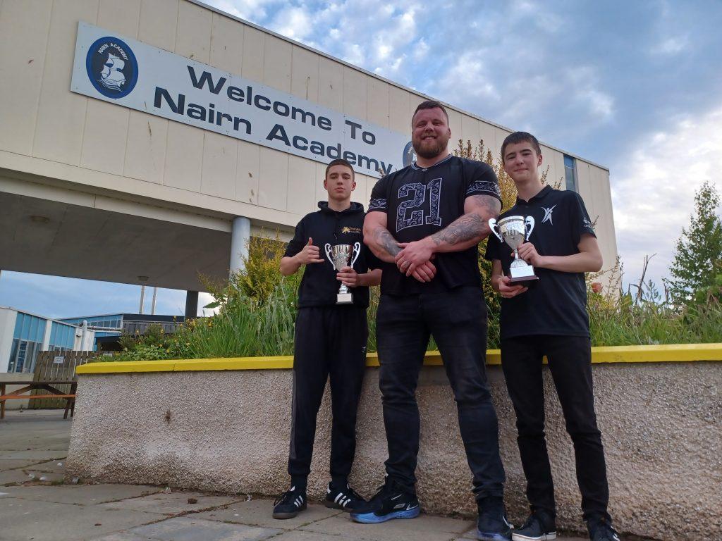 Three people standing in front of Nairn Academy; two are holding trophies.