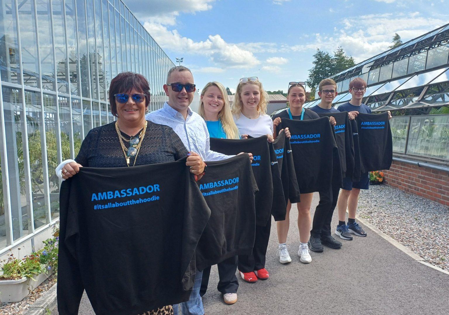 Seven people standing outside a greenhouse holding black "AMBASSADOR" hoodies with "#itsallaboutthehoodie" printed in blue.