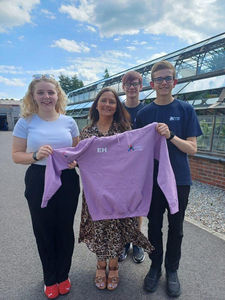 Four people outside a greenhouse, one holding a pink hoodie with initials "EH" and an emblem.