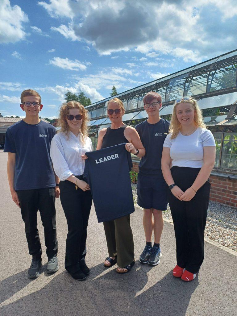 Five people outside a greenhouse, one holding a black "LEADER" hoodie.