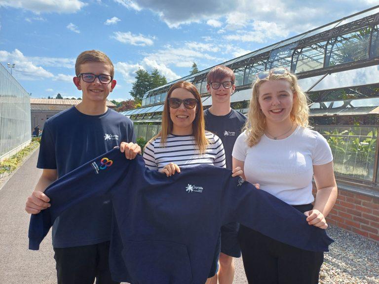 Four people outside a greenhouse, one holding a navy blue hoodie with an emblem.