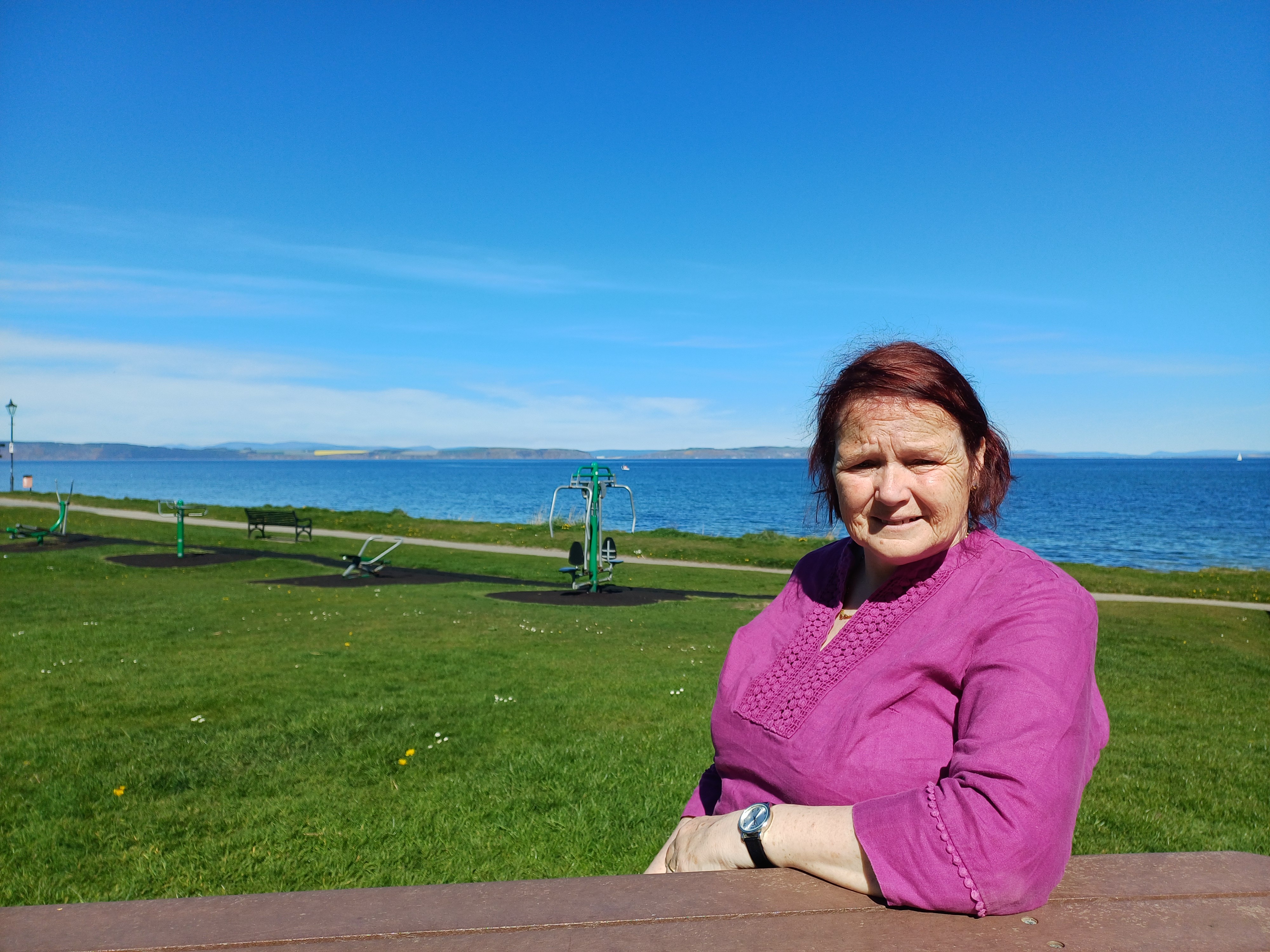Woman in pink top sitting at an outdoor bench with exercise equipment and the sea in the background on a sunny day