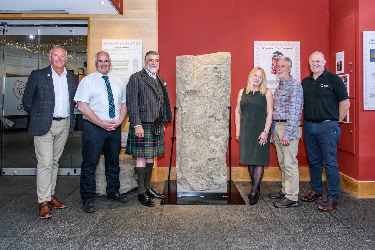 Six people standing around the Pictish stone in a museum exhibit.