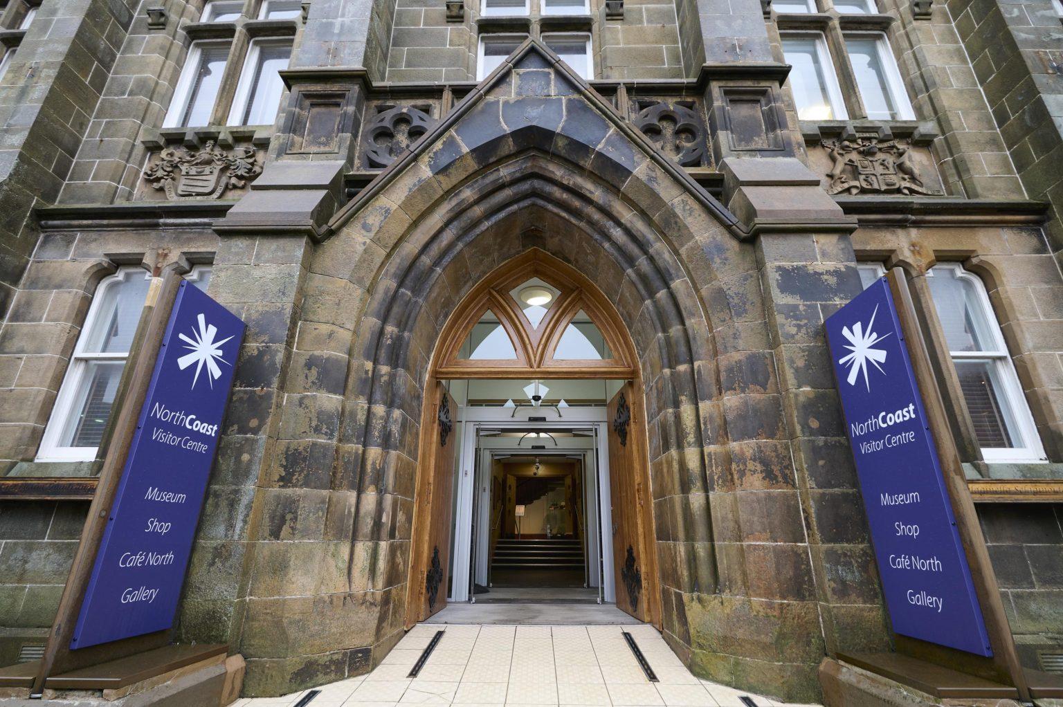 Entrance to the North Coast Visitor Centre, a stone building with a large arched doorway and blue signage indicating a museum, shop, café, and gallery.