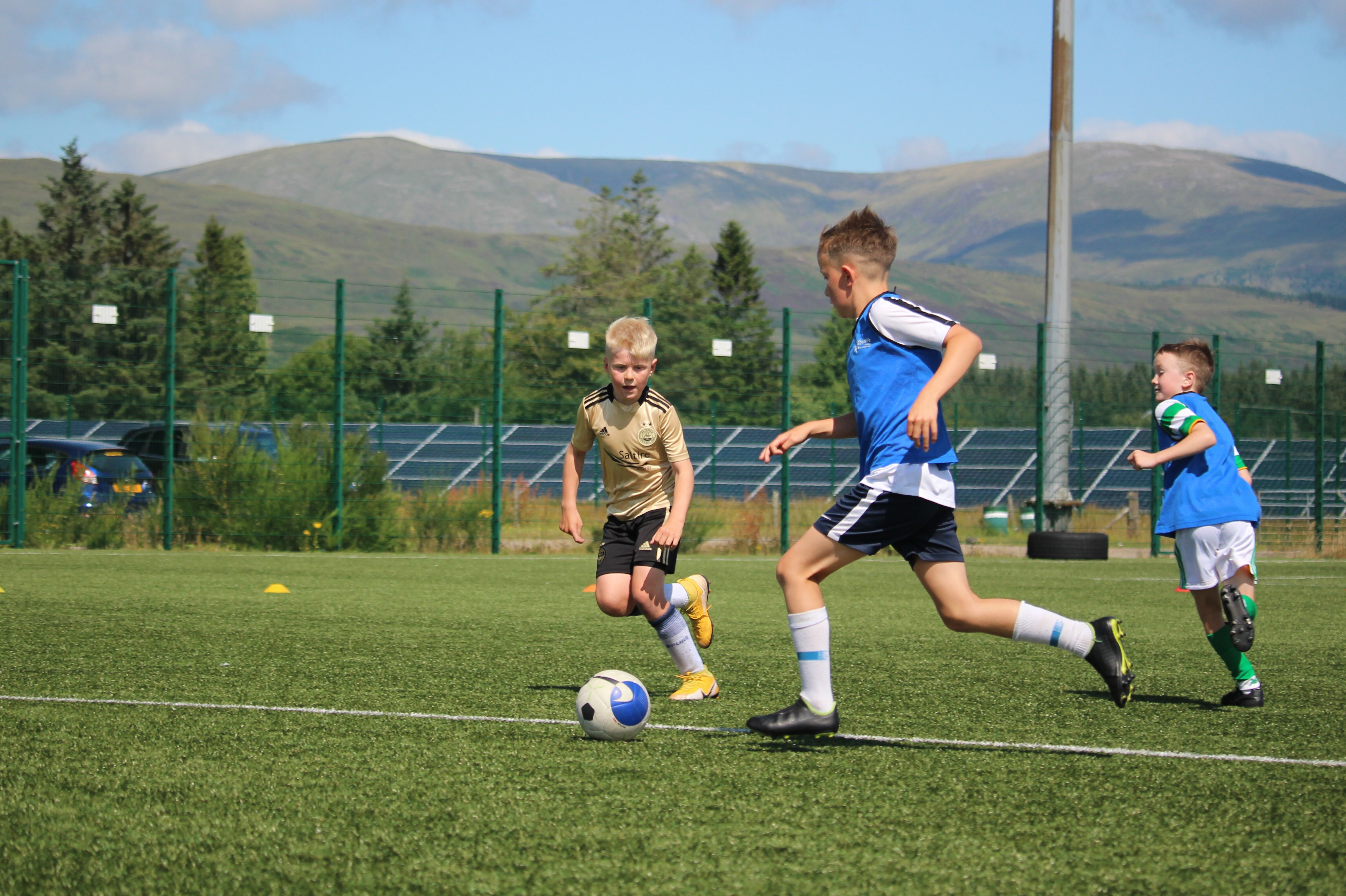 Three boys chase a football on an outdoor pitch with Highland hills and blue sky in the background