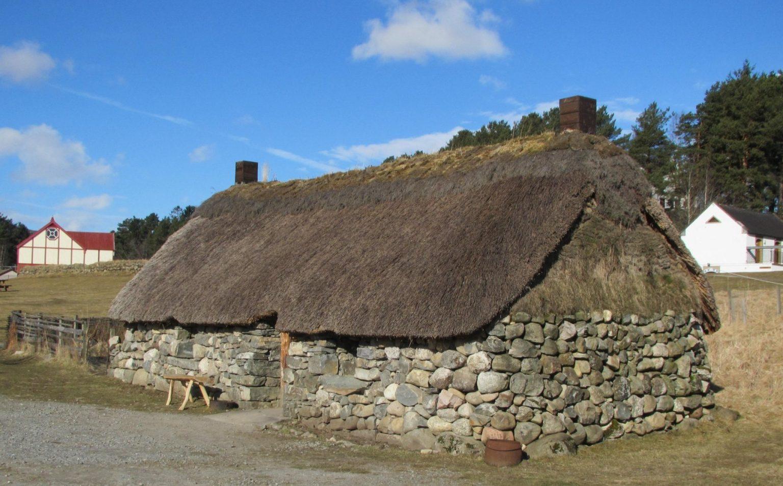 Traditional stone-built cottage with a thick thatched roof, located in an open outdoor setting with grassy ground and wooden fencing. Two chimneys rise from the roof, and there are trees and additional buildings in the background under a bright blue sky with scattered clouds.