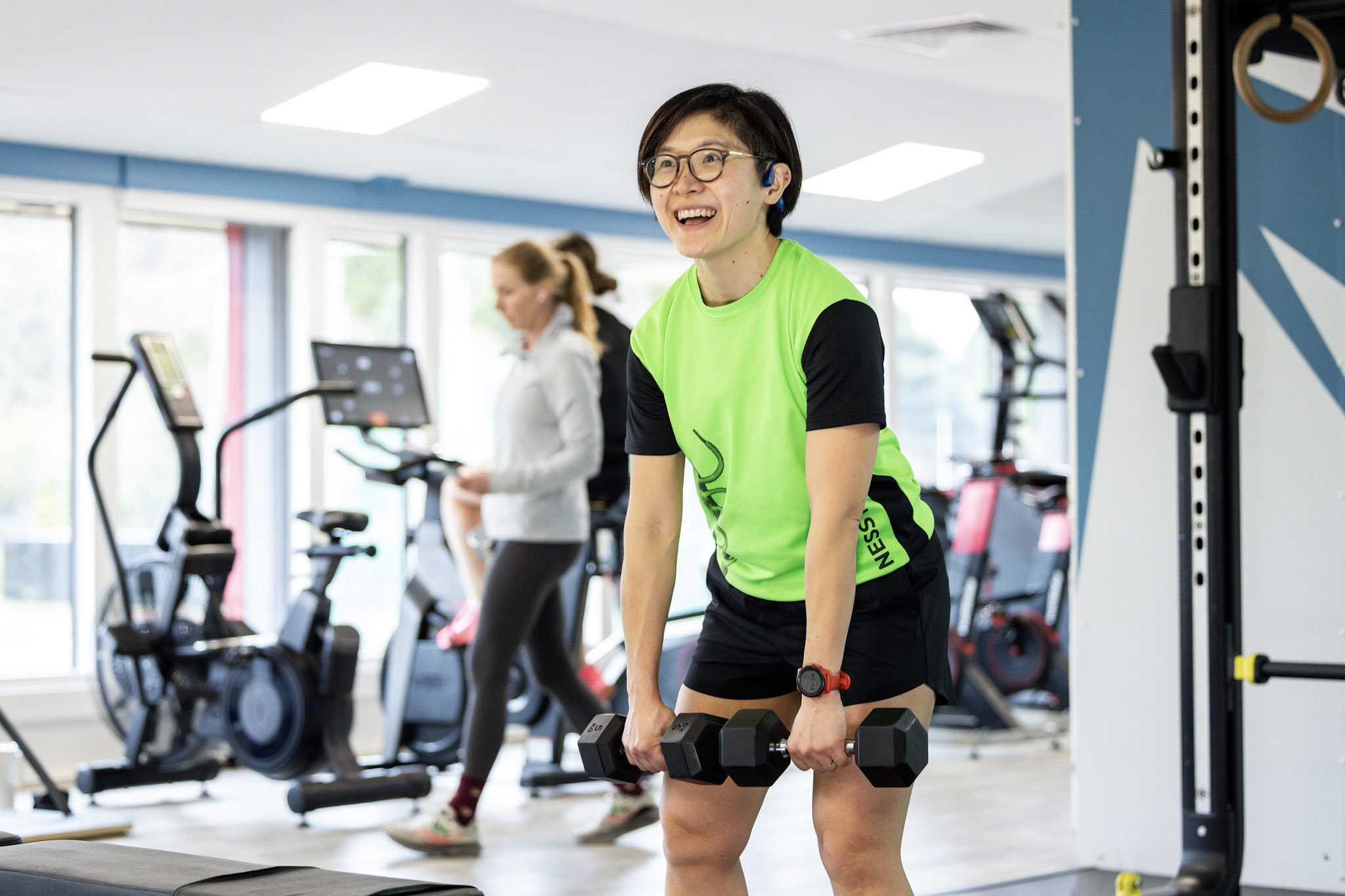 A woman smiles while lifting dumbbells in a bright, modern gym