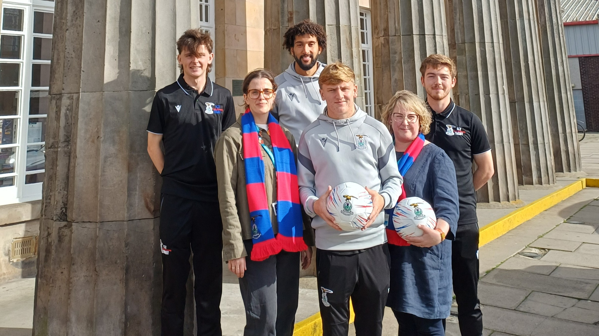 Two Inverness Caledonian Thistle players, two ICT Community Trust coaches and two library staff stand outside Inverness Library holding club footballs and scarves.