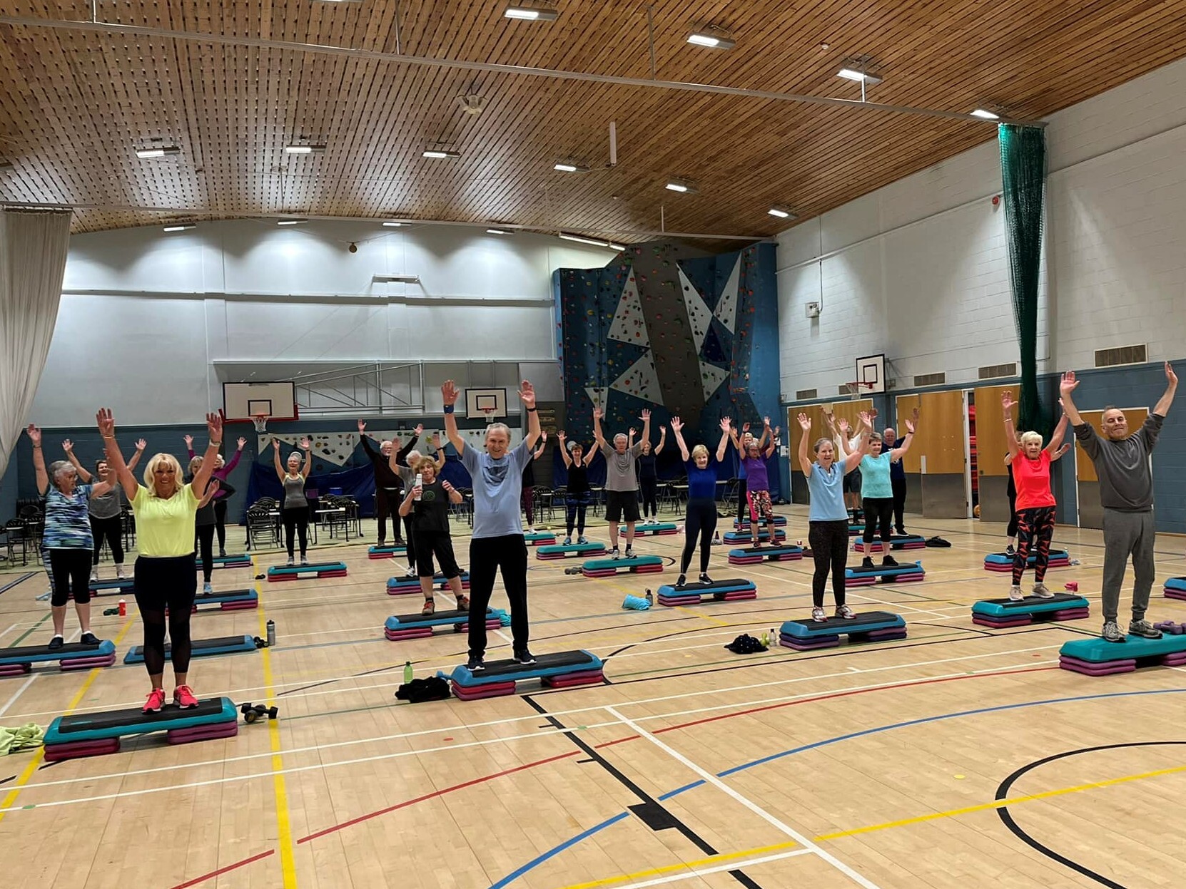 A large group of older adults taking part in a Fit Plus exercise class in a sports hall, all with arms raised above their heads, step platforms in front of them.