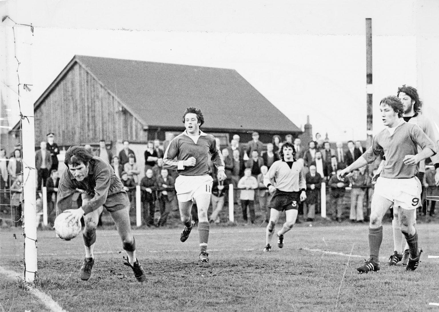 Black and white photo of a football match showing a goalkeeper near the goalpost bending to pick up the ball, with players wearing numbers 11 and 9 nearby. A wooden building and a crowd of spectators behind a fence are visible in the background.