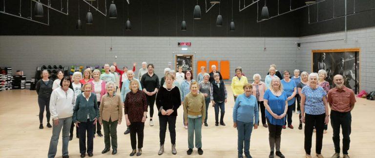 Group of people standing in a gym or fitness studio with wooden floors, mirrors, and exercise equipment.