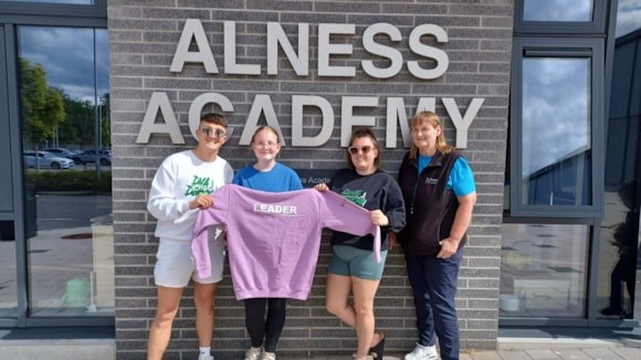 Four people standing outside Alness Academy holding a purple sweatshirt with the word "LEADER" on it.