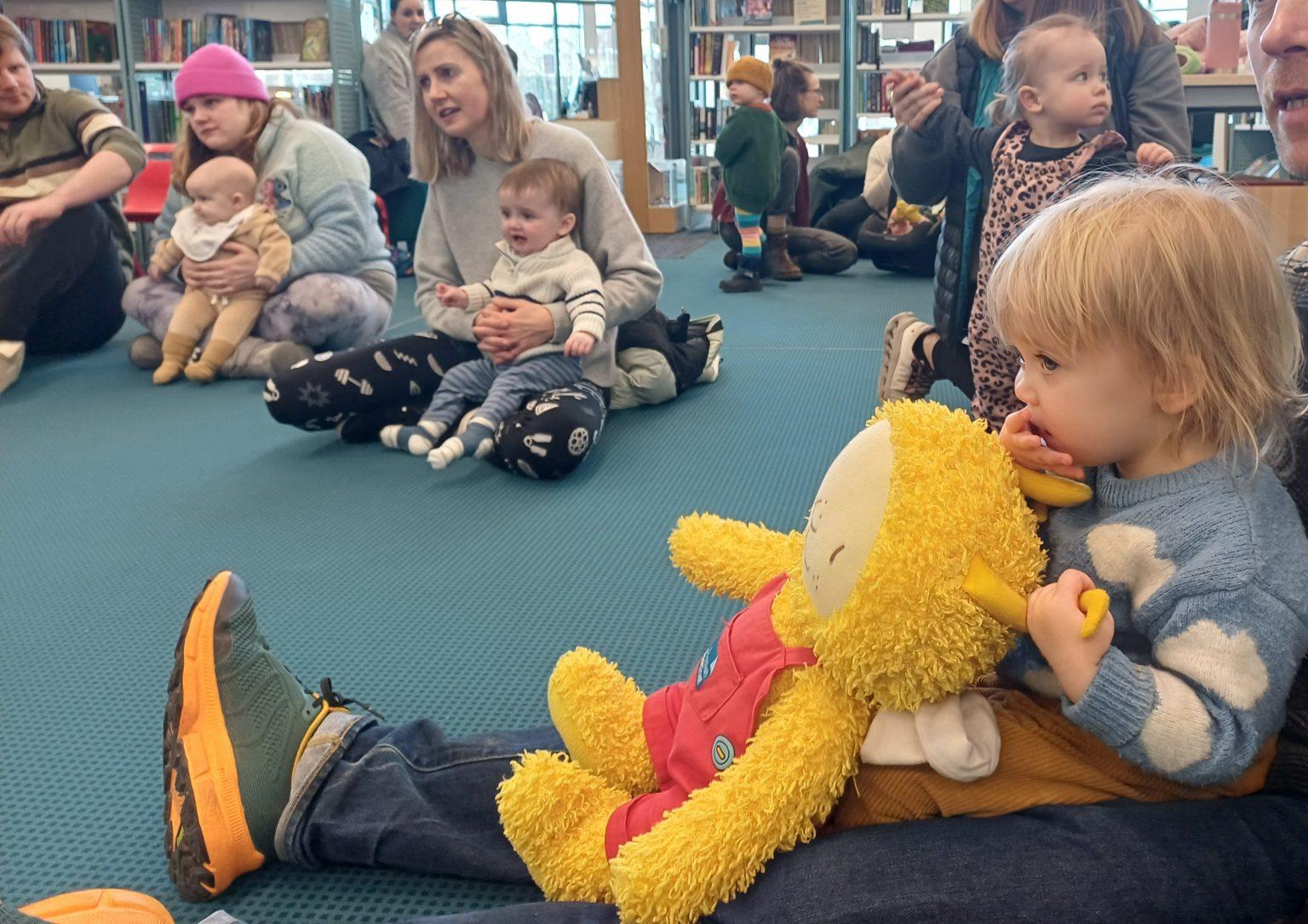 Children and parents seated on a blue carpeted floor during a Bookbug session in a library, surrounded by bookshelves. The children hold soft toys while adults engage with them in a relaxed, interactive setting.