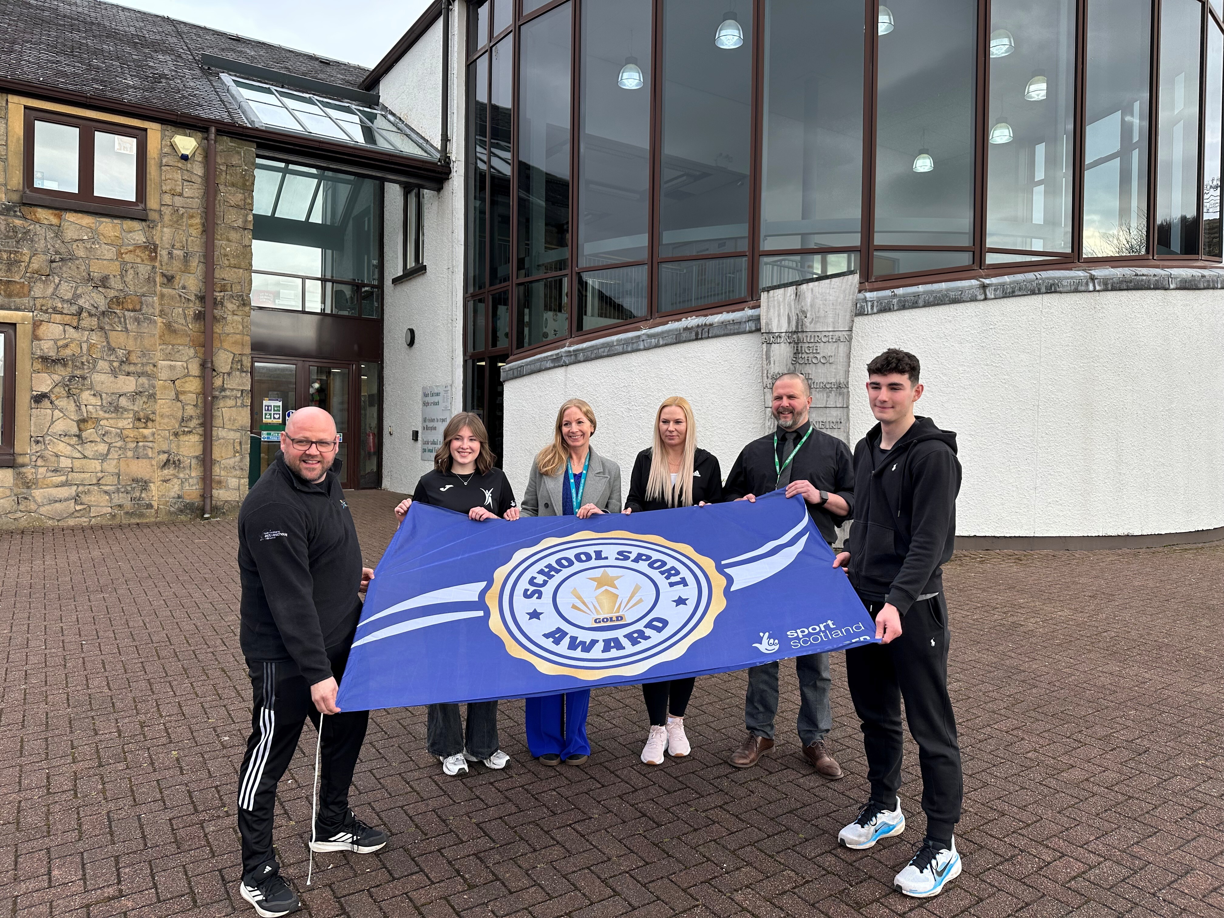 A group of six people stands outside a school building holding a large blue flag that reads ‘School Sport Award Gold.’ The group is gathered on a paved area near the building’s entrance.