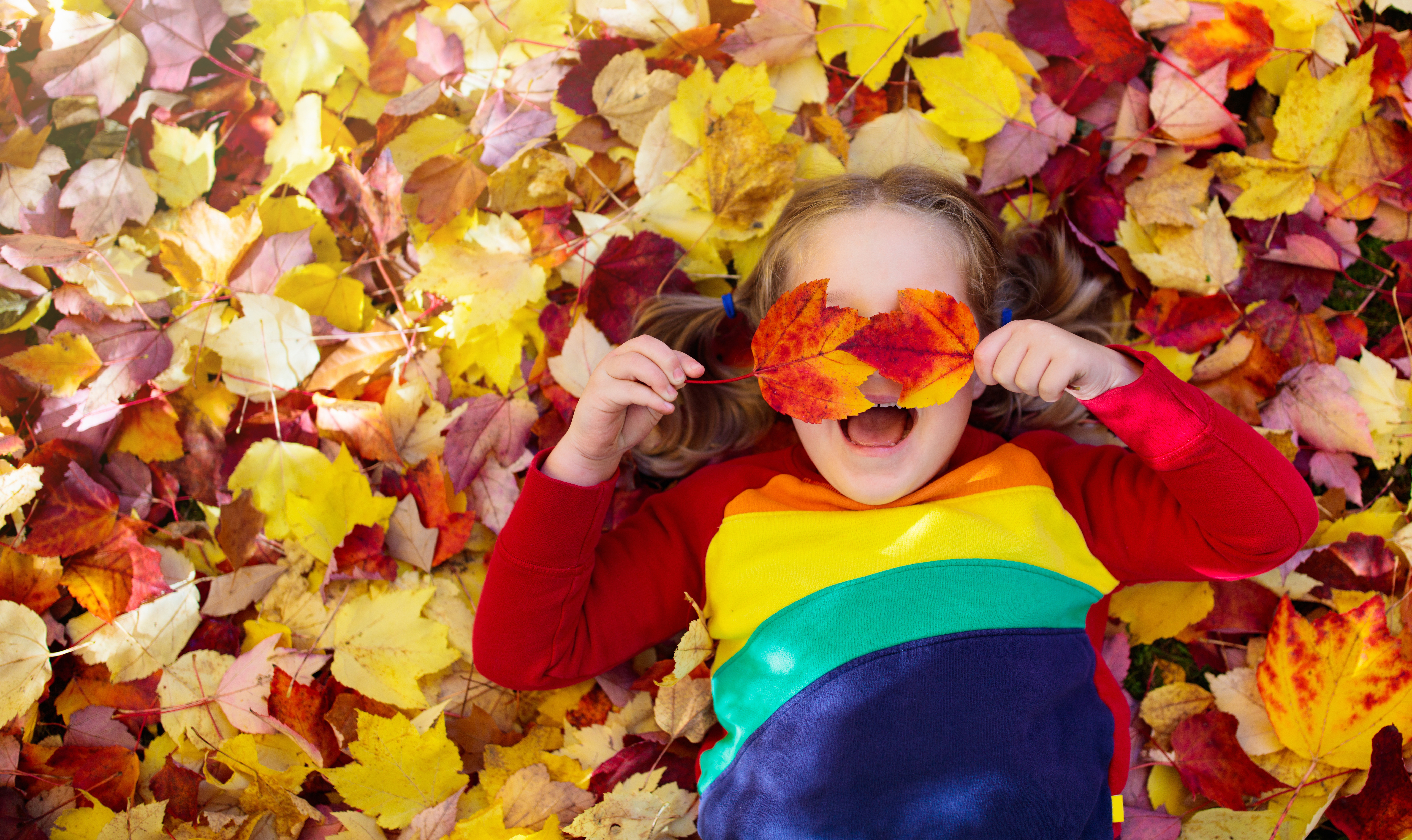 A child lies in a pile of colourful autumn leaves, laughing and holding two red leaves over their eyes.