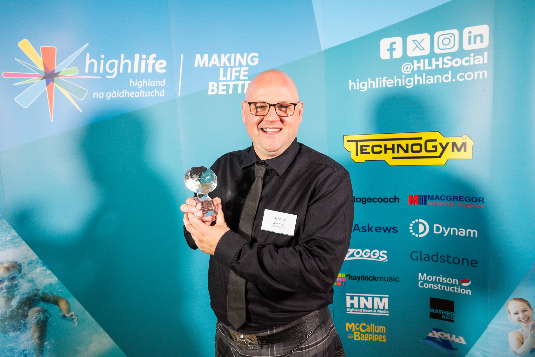 A man in a dark shirt and tie and kilt, with glasses. He is standing in front of a blue sponsorship board holding a small glass trophy.
