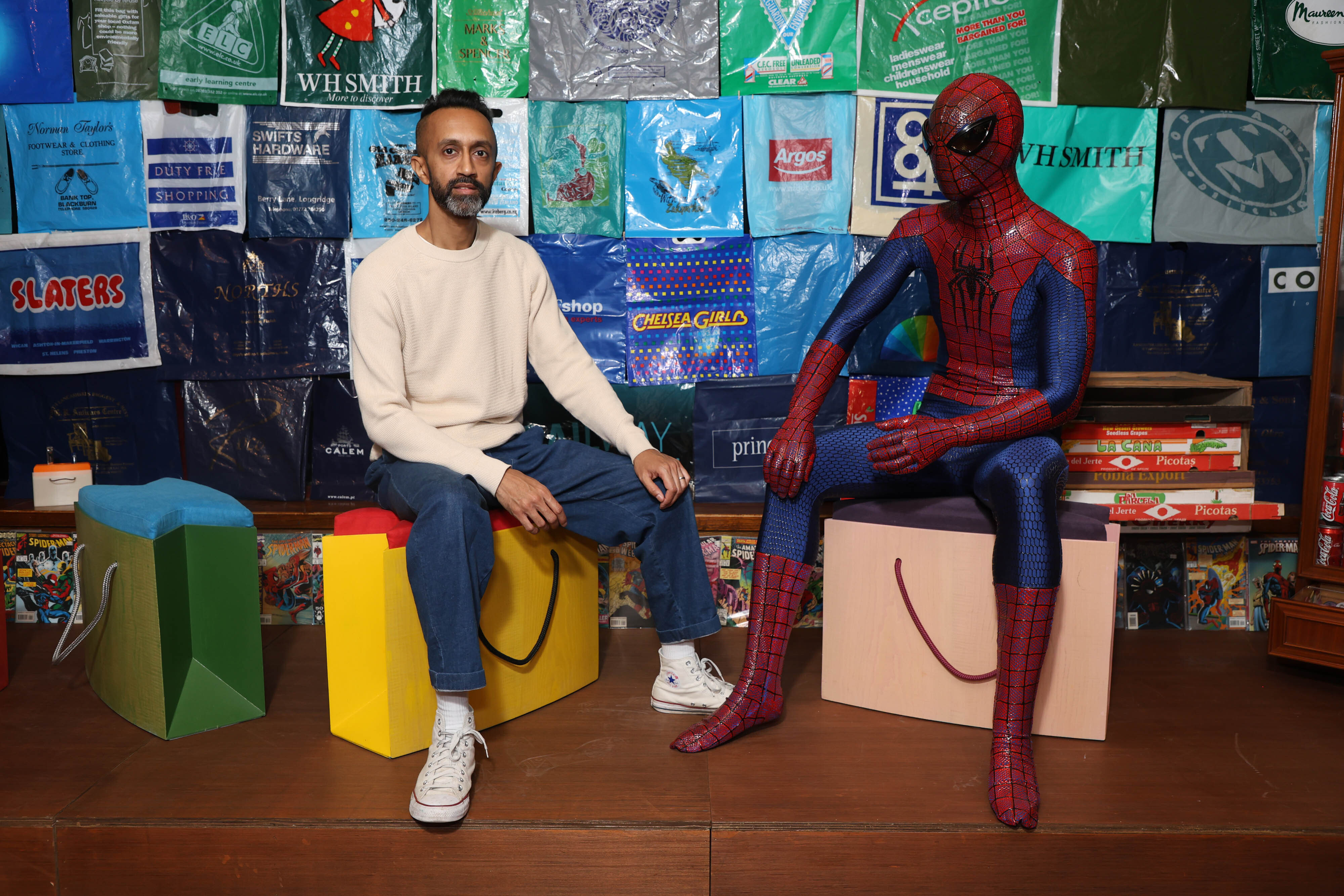 A man in a white jumper and blue jeans sits on a yellow seat in front of a collection of bags alongside him is a man sitting on a pink seat dressed as Spiderman.