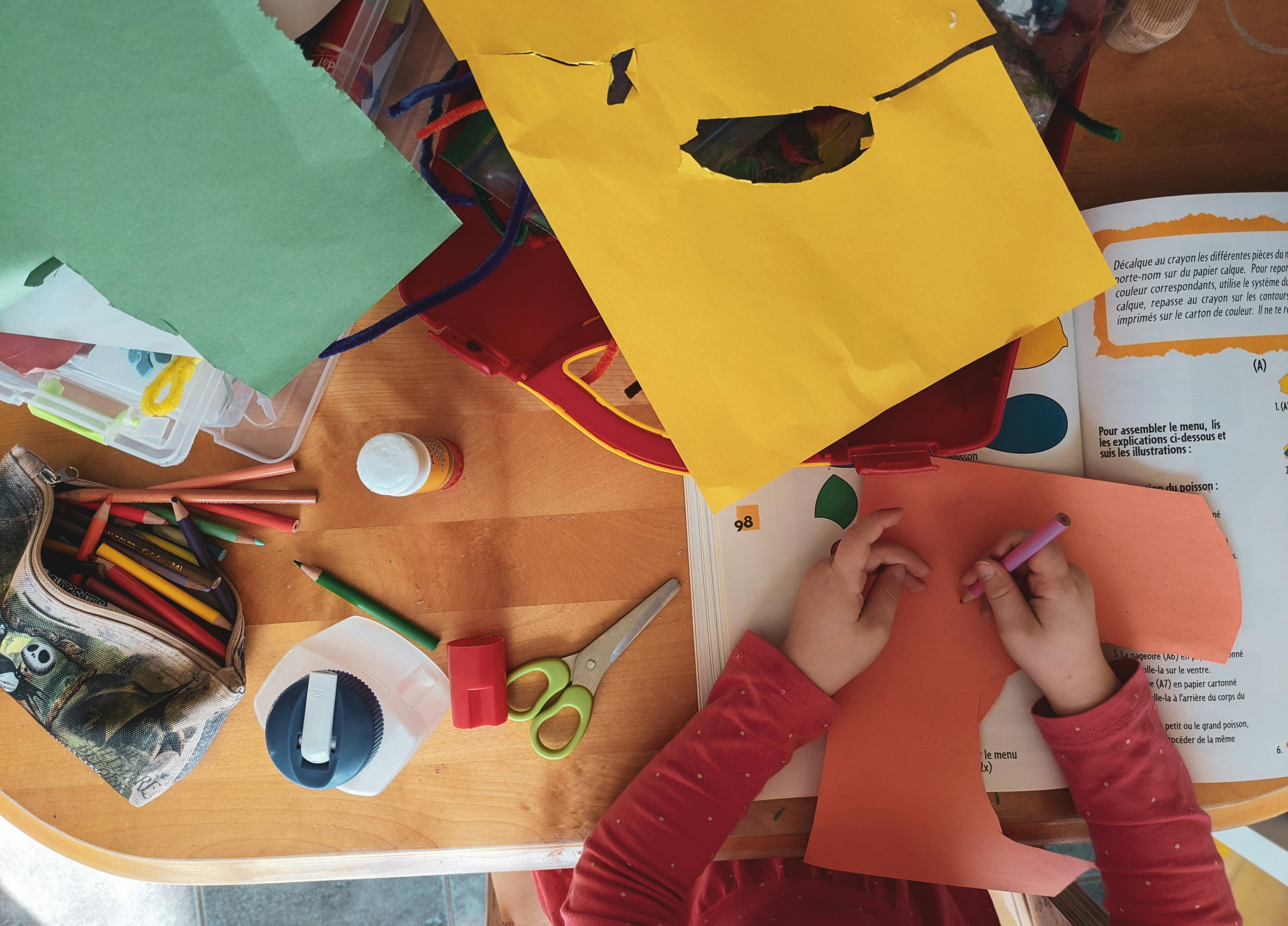 A child's hands colour on orange paper at a table covered in craft supplies, including coloured pencils, scissors and glue.