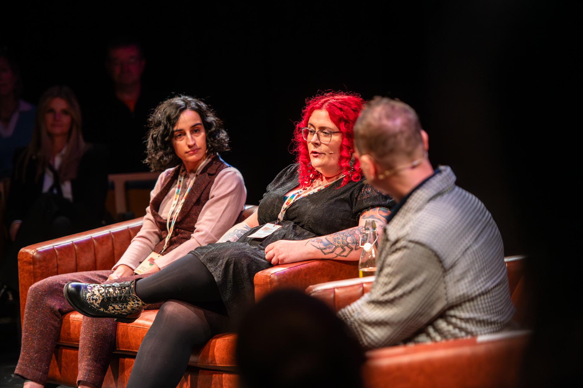 A woman with dark hair is sitting on a brown sofa. Sitting to her right is a woman with red hair who is talking. There is a man to her right with his back to the camera. They are in a theatre setting.