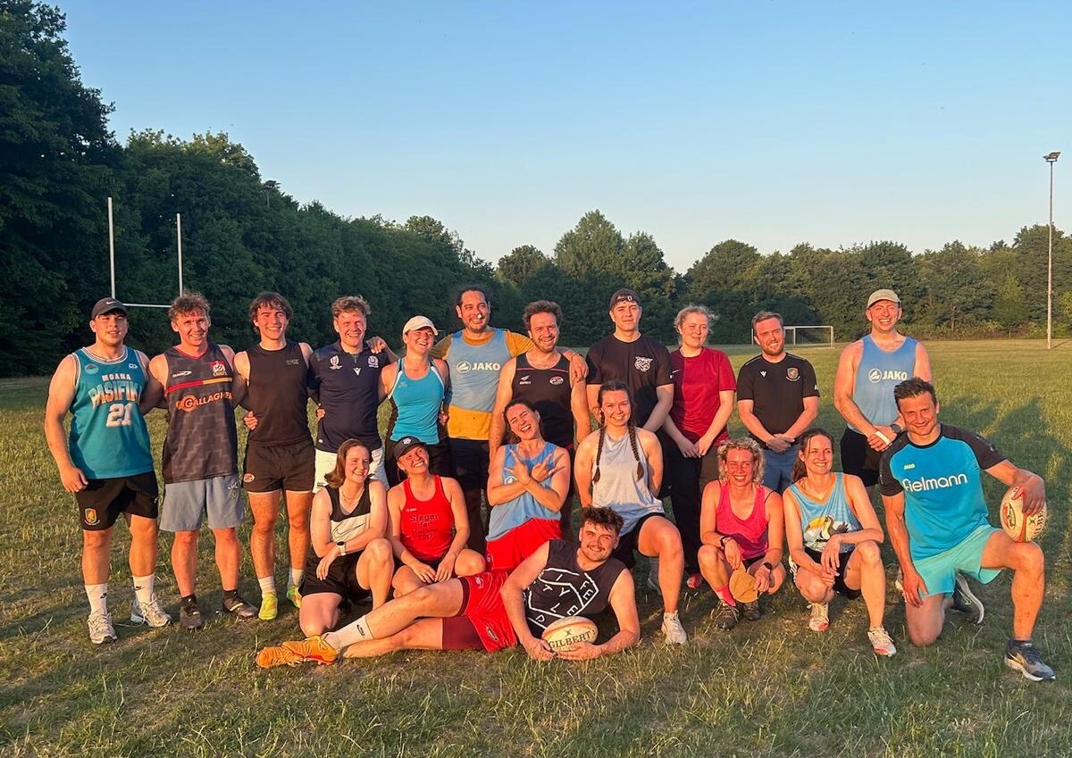 Around 20 people are gathered together for a photo in the sunshine. They are wearing sports gear and have just taken part in an informal game of rugby.