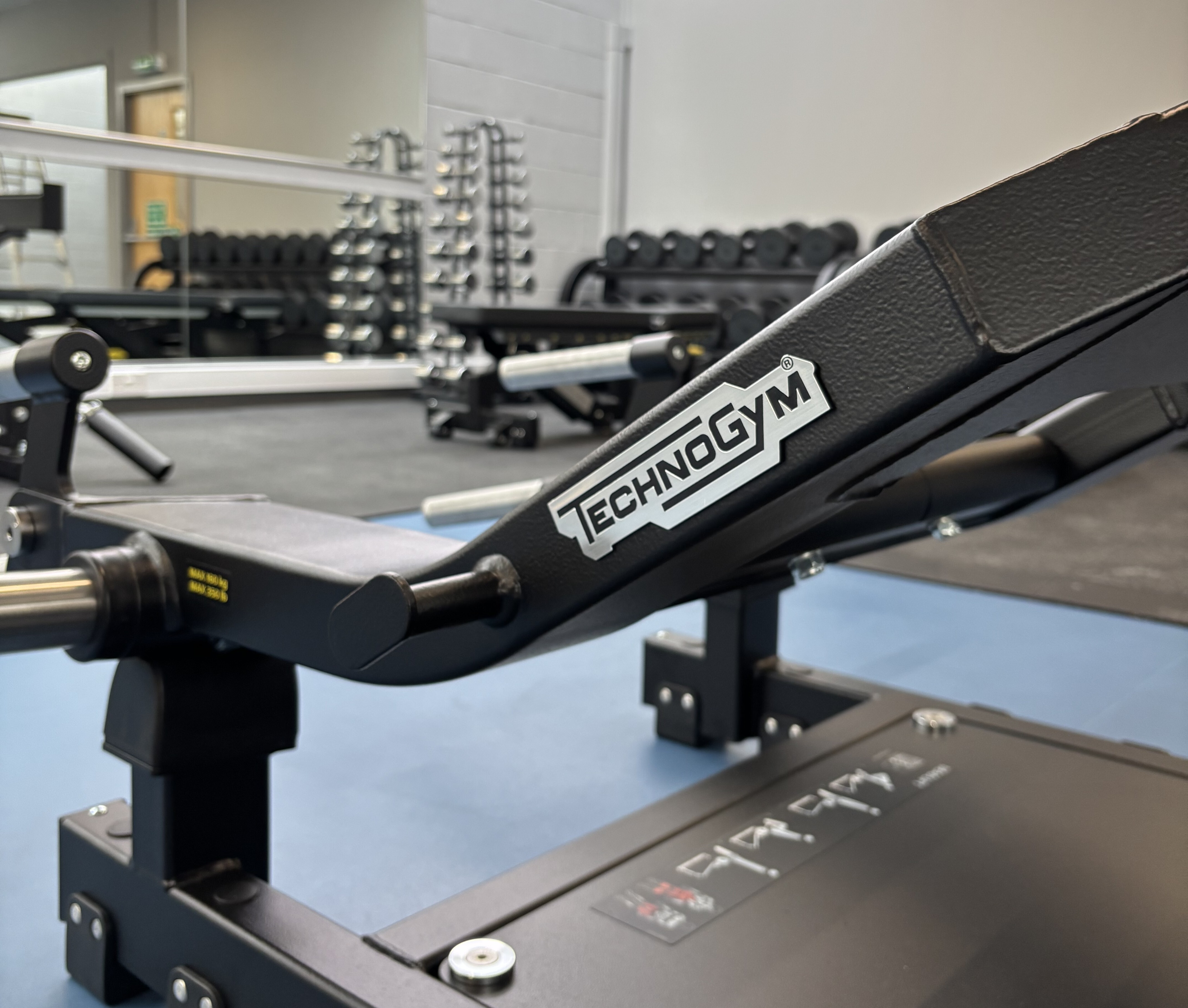 A close up of a piece of Technogym gym equipment. There are weights and other gym equipment in the background.