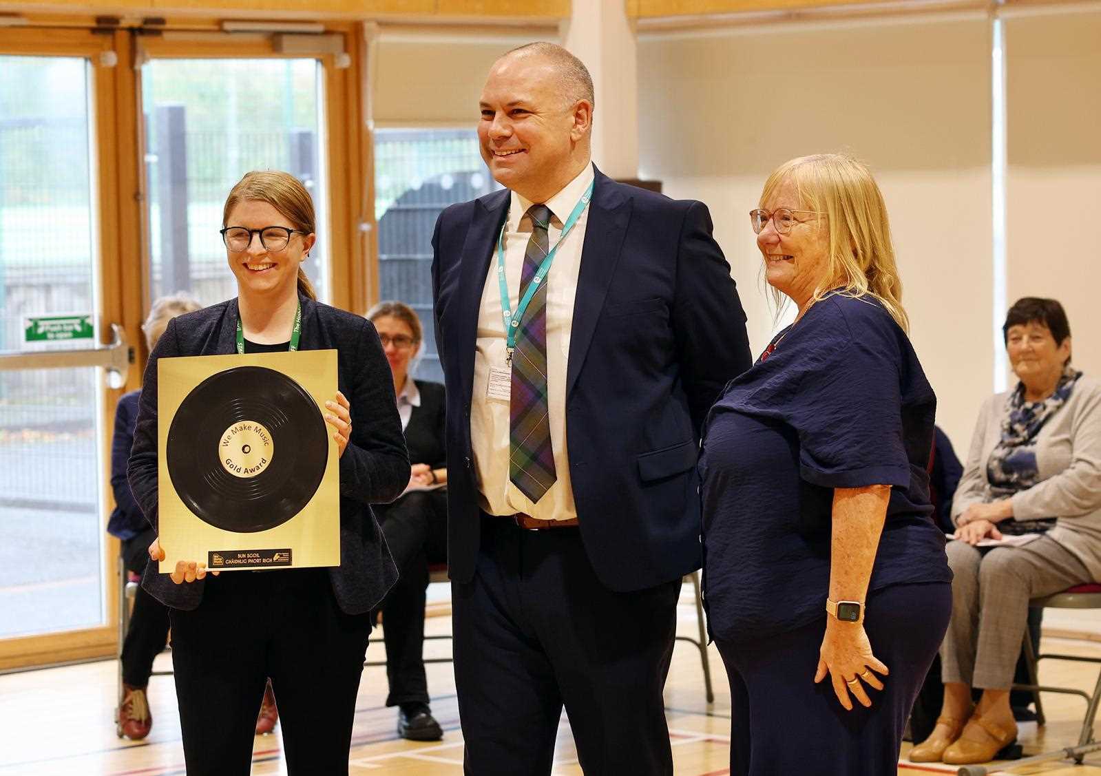 A male in a suit and tie with two females either side of him. The female on the left is holding an award as the female on the right look son.
