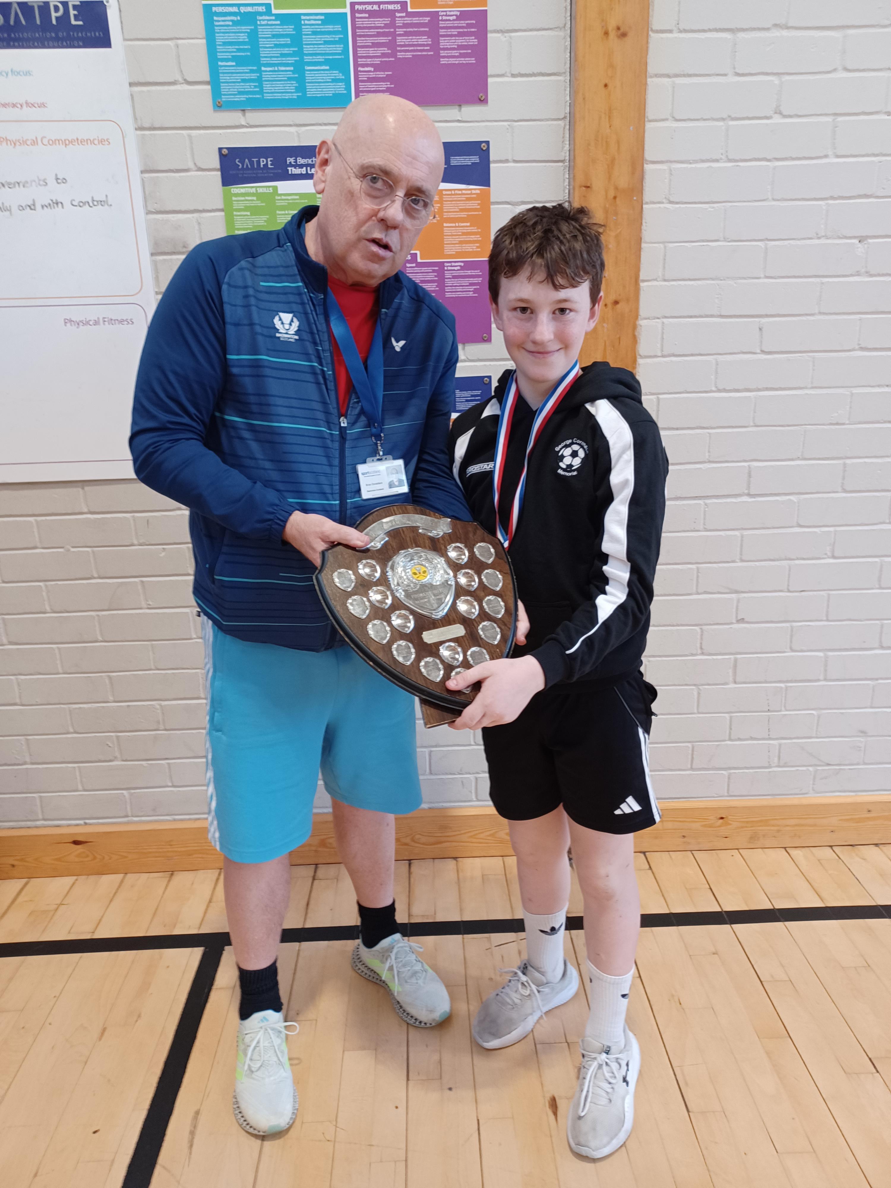 A young boy holding a shield trophy alongside an adult at the East Sutherland Schools badminton tournament at Golspie High School.