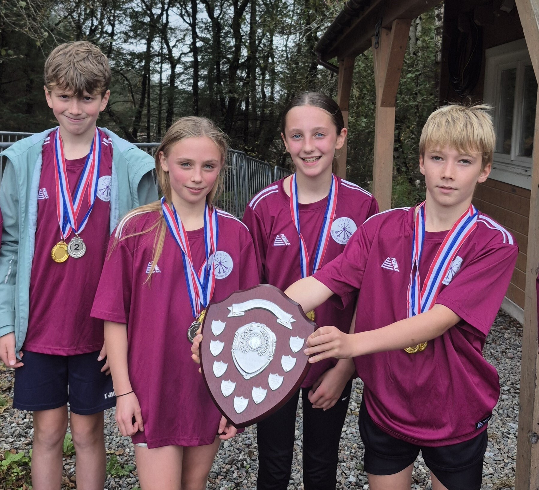 Four young people - two boys and two girls - wearing purple tops and sports wear are holding up a winners shield and wearing medals. They are standing outside a cabin.