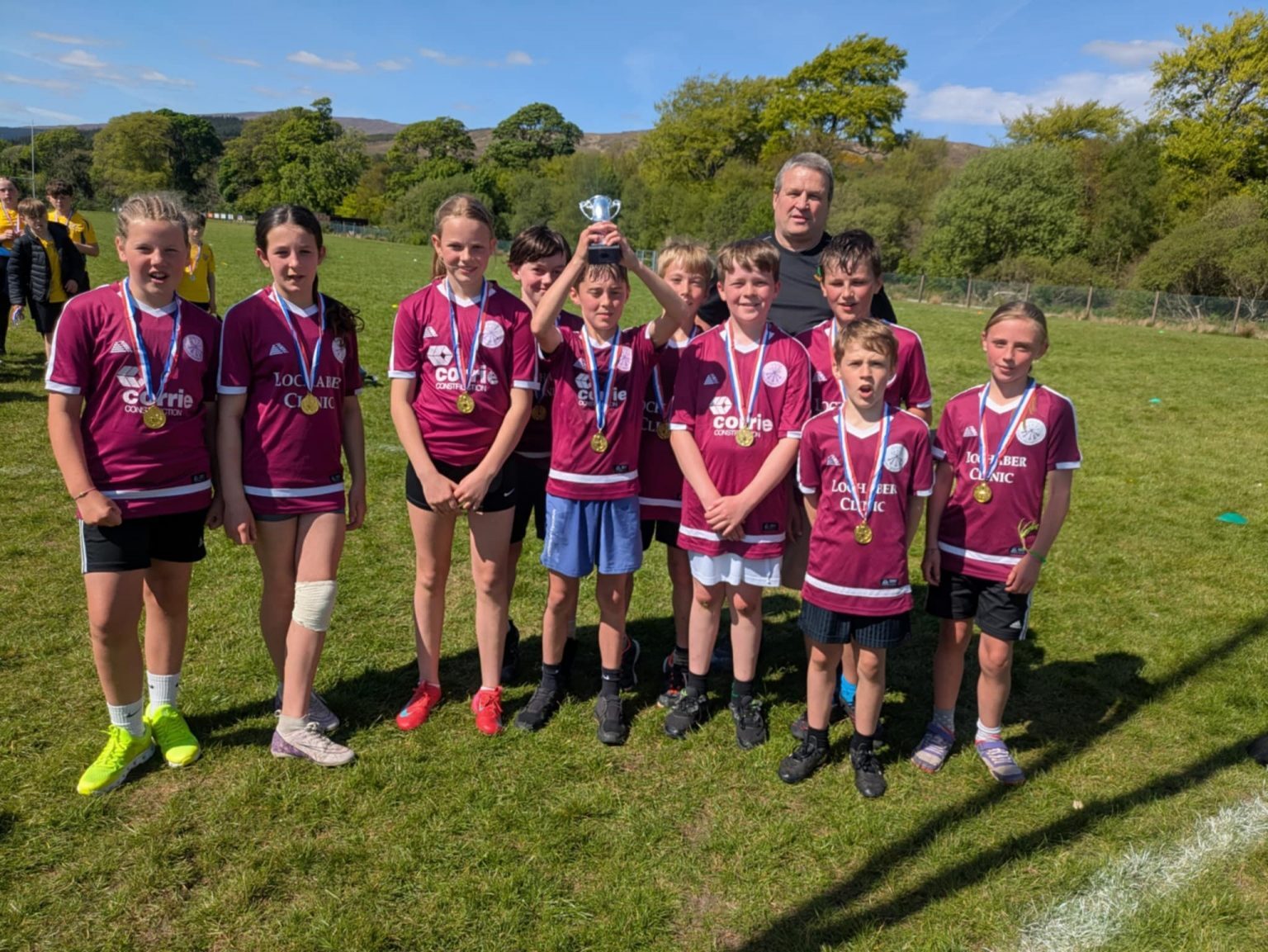 A group of young athletes wearing matching maroon sports jerseys with white trim and black shorts stand together on a grassy field. Each player has a medal around their neck, and one person in the center is holding a small silver trophy above their head.