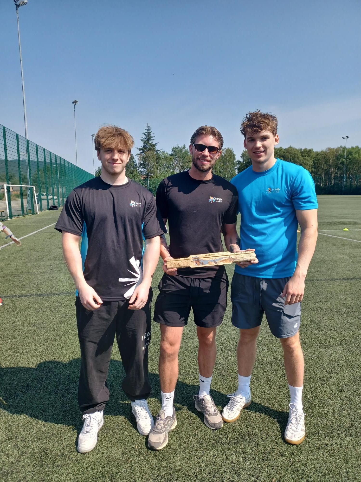 Three young High Life Highland staff holding the King's Baton on a sports pitch