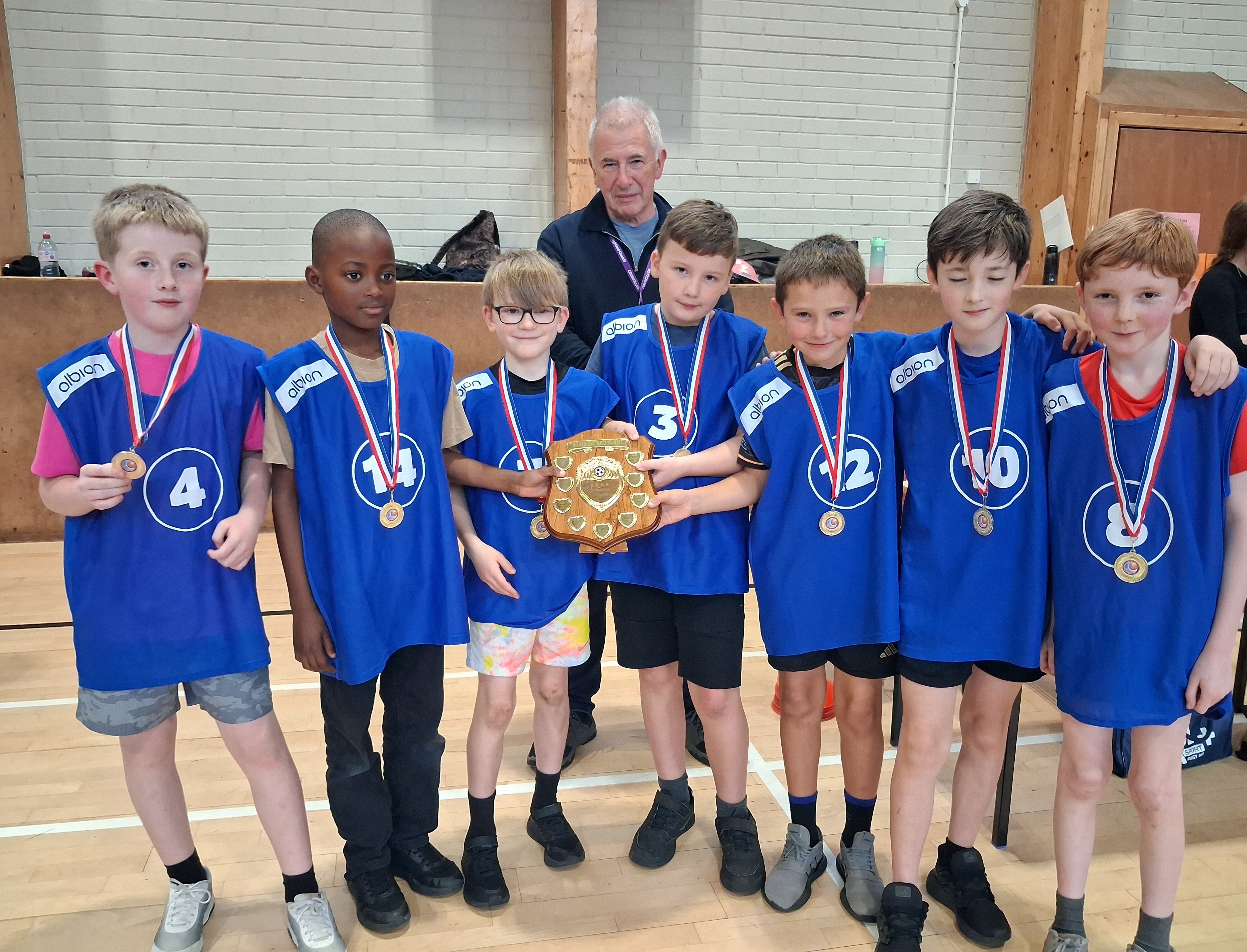 A group of primary school boys in blue bibs and medals hold a trophy shield in a sports hall. An older man stands behind them.