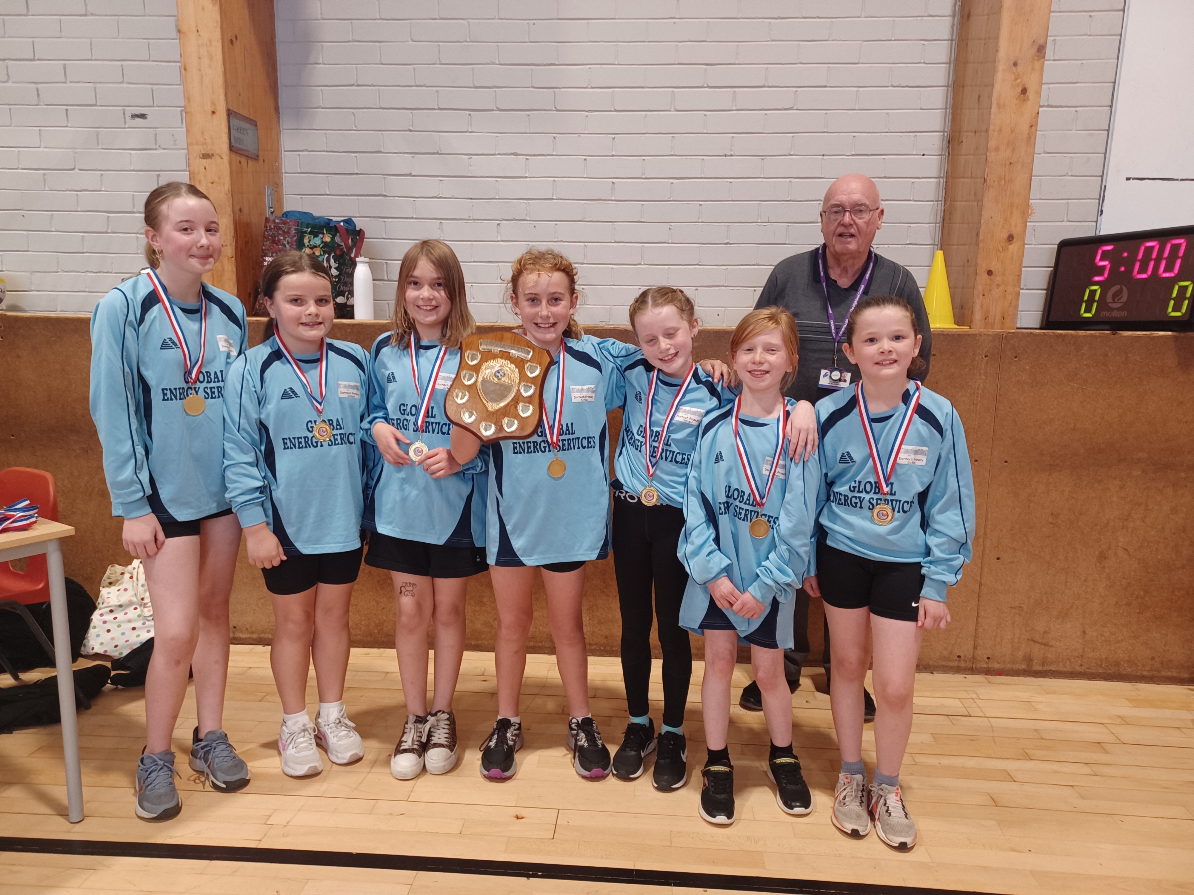 A group of seven young girls in pale blue tops and sportswear. They are in a sports hall and are holding up a winners shield.