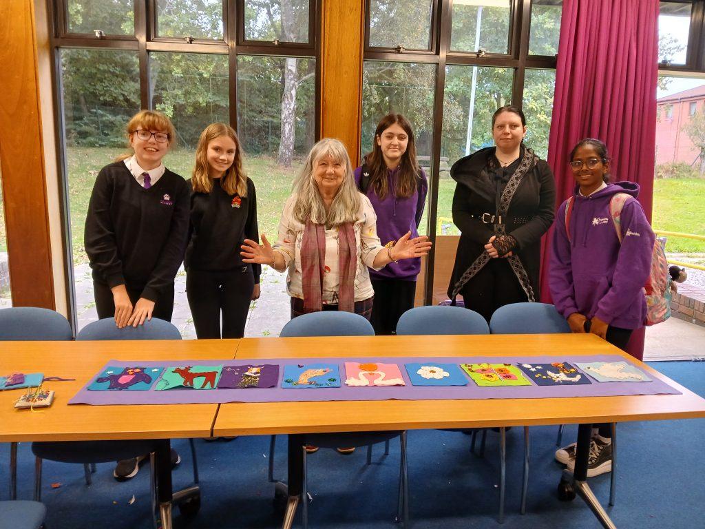Six people stand behind a table displaying colorful fabric artworks featuring animals, flowers, and abstract designs at Culloden Library.