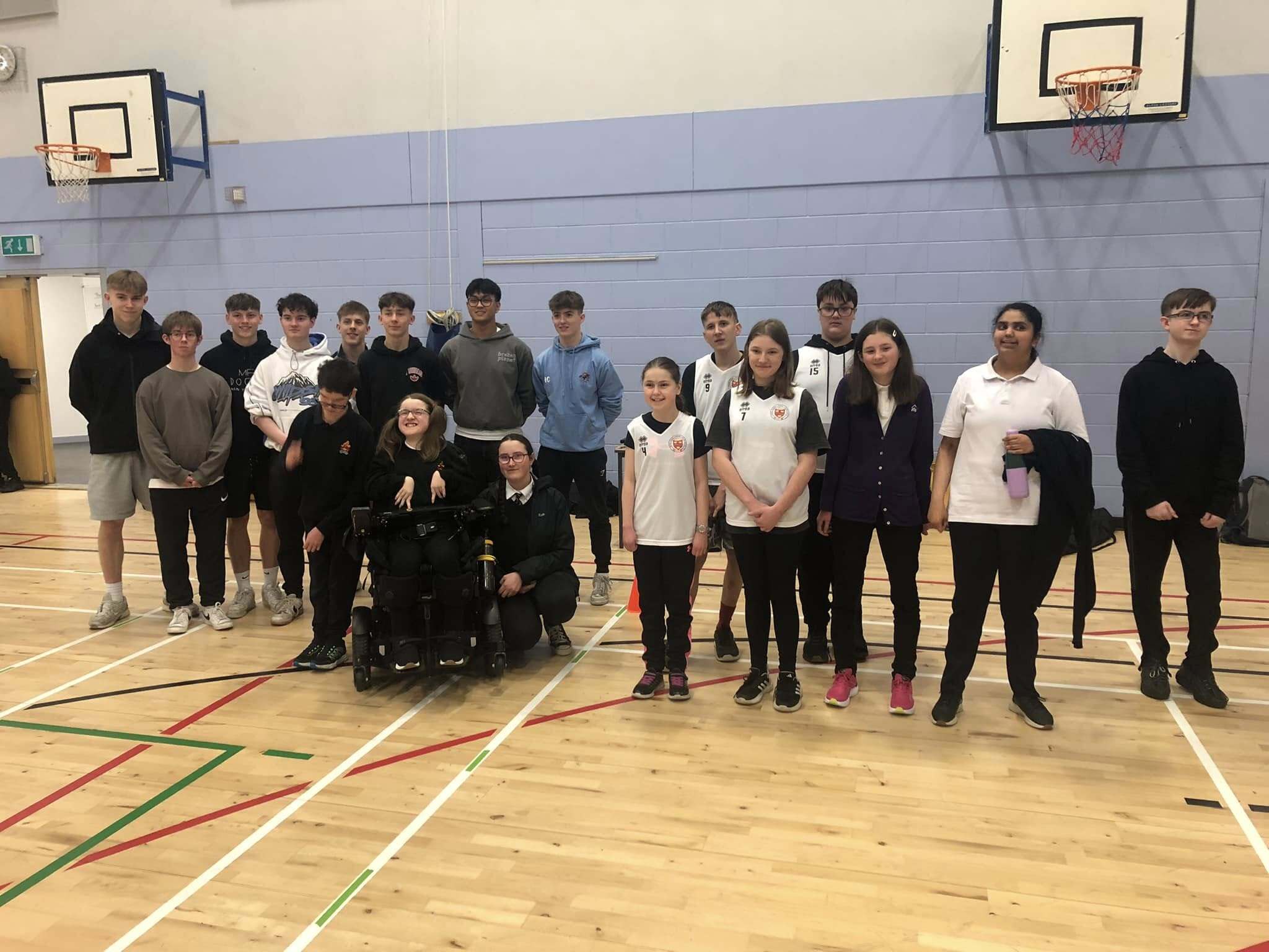 A large group of high school pupils of various ages are gathered together in a sportshall. One pupil is in a wheelchair.