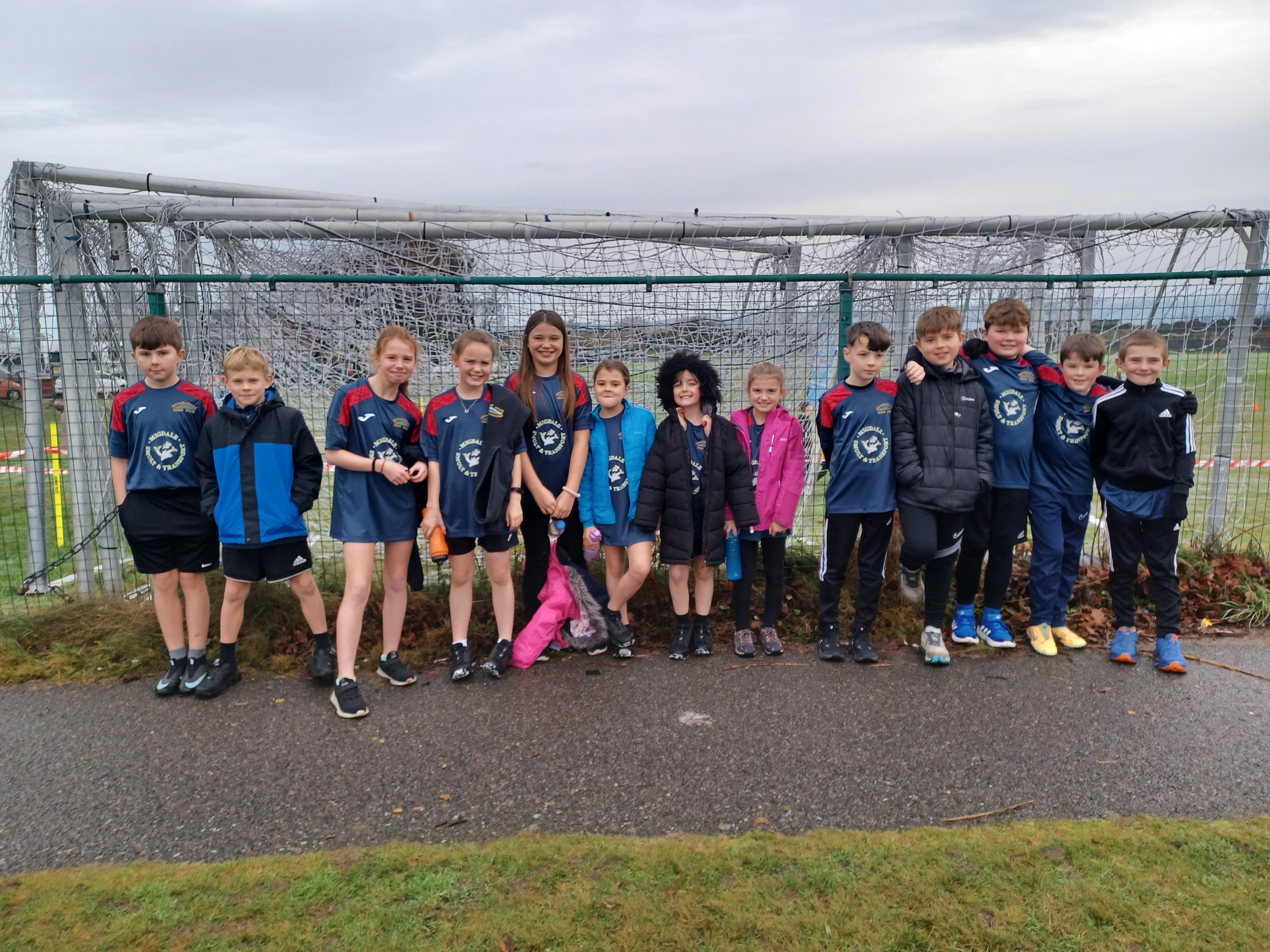 13 school pupils lined up against a sports pitch fence.