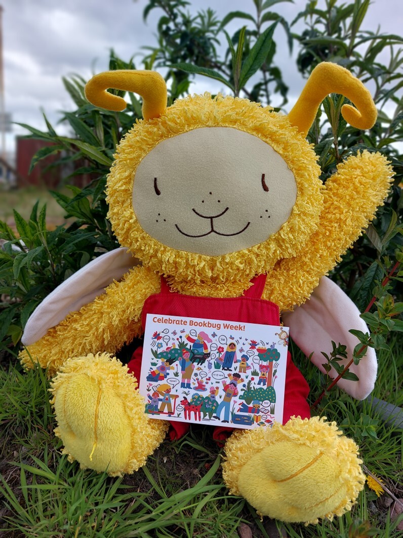 A plush yellow character with antennae and white wings is sitting on green grass in front of leafy plants. The character is wearing a red outfit and holding a colorful sign that reads “Celebrate Bookbug Week!”